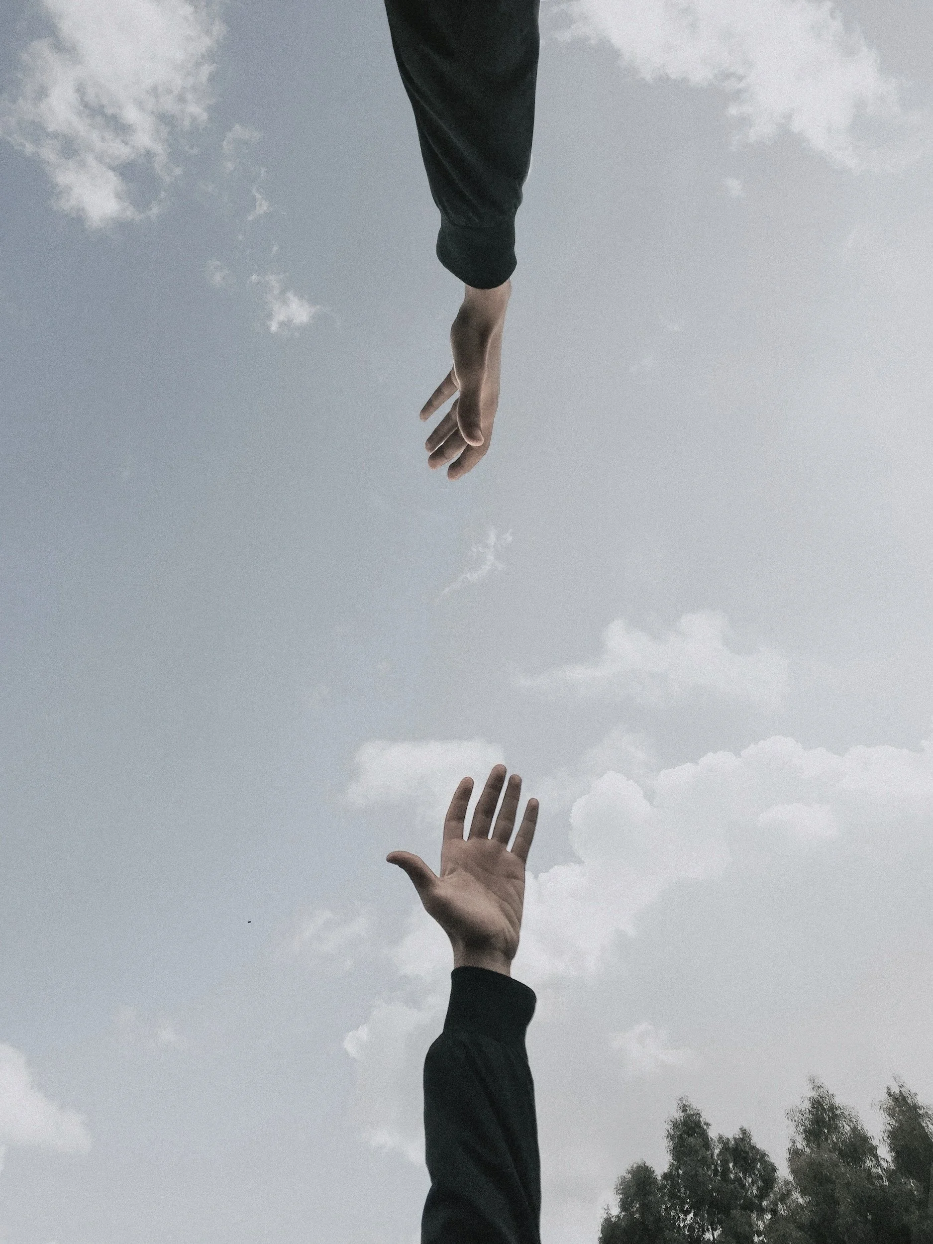 Two hands reaching towards each other against a cloudy sky, one higher and one lower, with trees in the background.