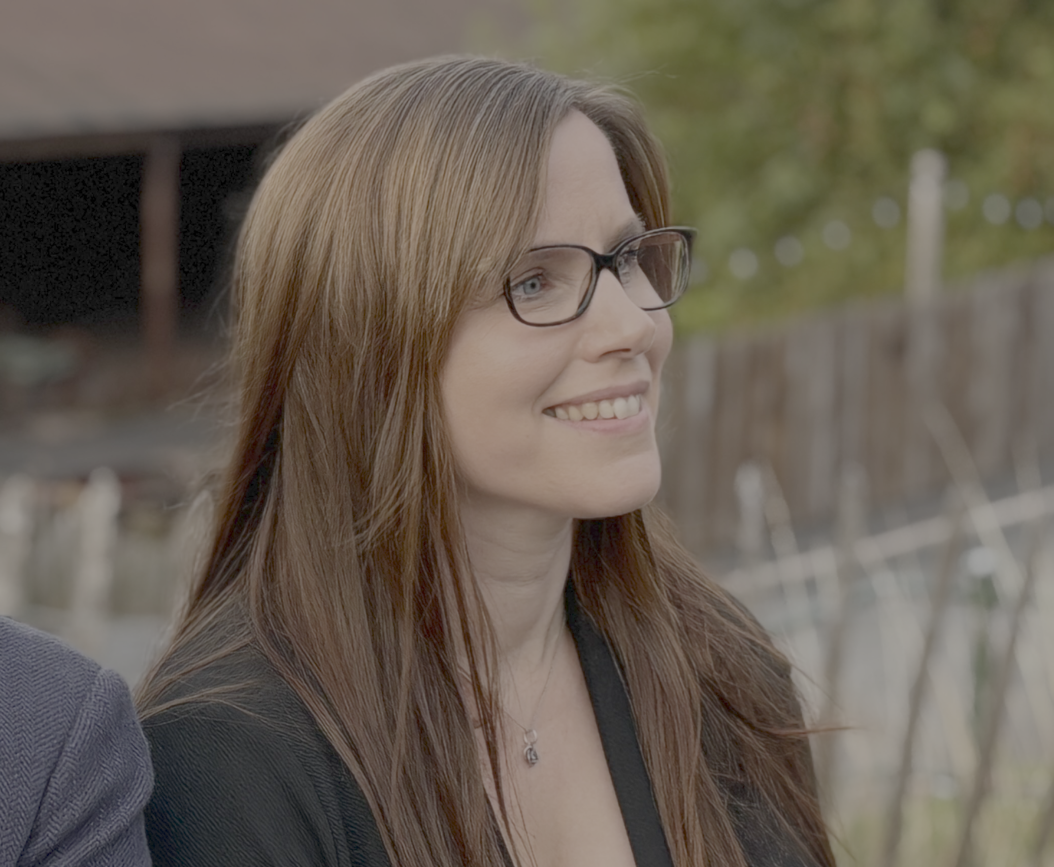 Rebecca Williams, wearing glasses and a black blazer, smiling outdoors with trees and a wooden fence in the background.