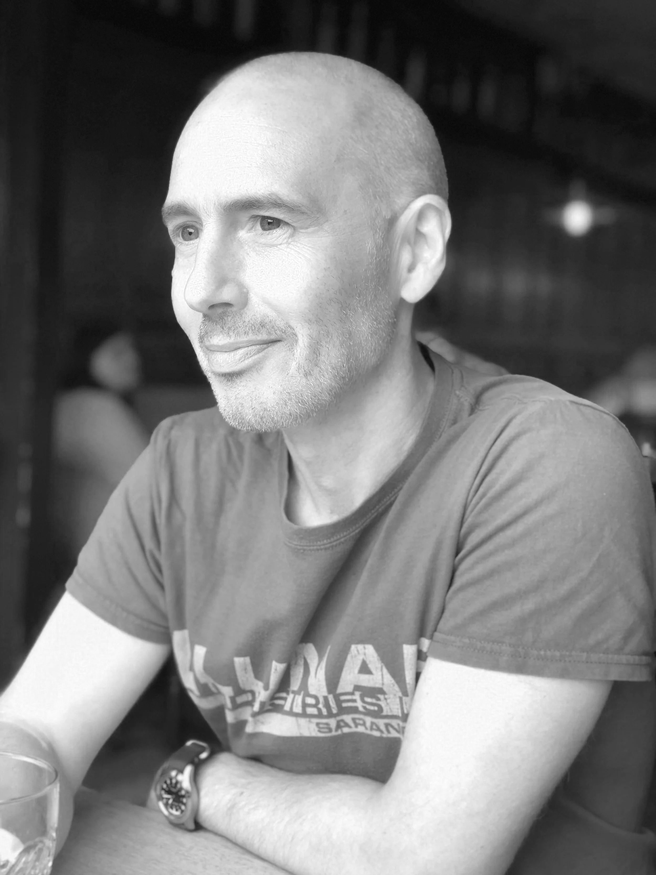 A man with a shaved head and a beard, wearing a t-shirt, sitting at a table with his arms crossed, smiling slightly, in a dimly lit indoor setting.