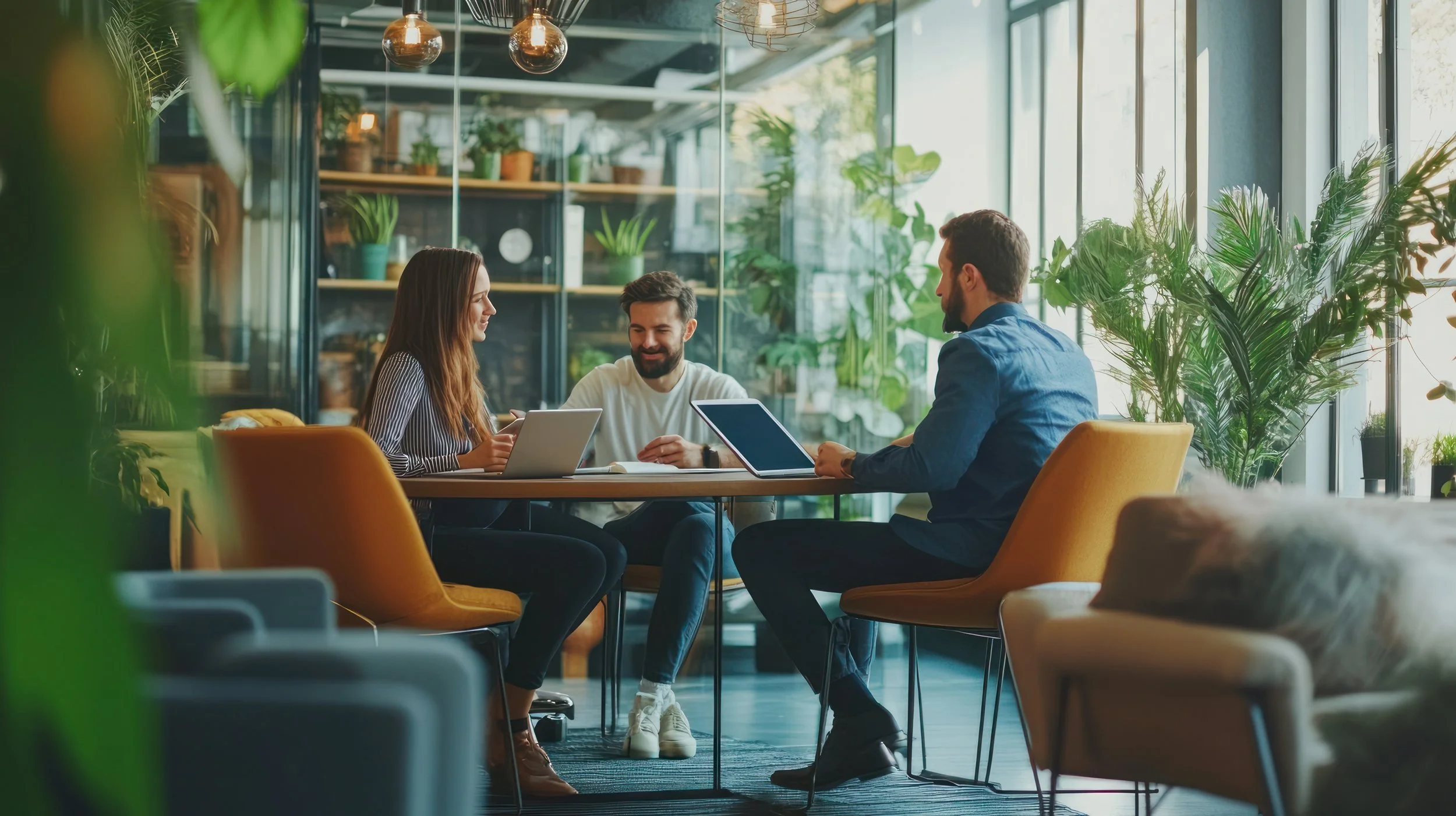 Trois consultants travaillent autour d'une table dans un espace de coworking moderne, entouré de plantes vertes et avec de grandes fenêtres laissant entrer beaucoup de lumière naturelle. Ils pensent innovation.