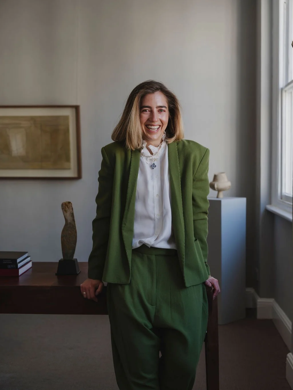 Portrait of a smiling woman with shoulder-length hair, wearing a suit jacket, a ruffled blouse, and layered necklaces, against a plain background.
