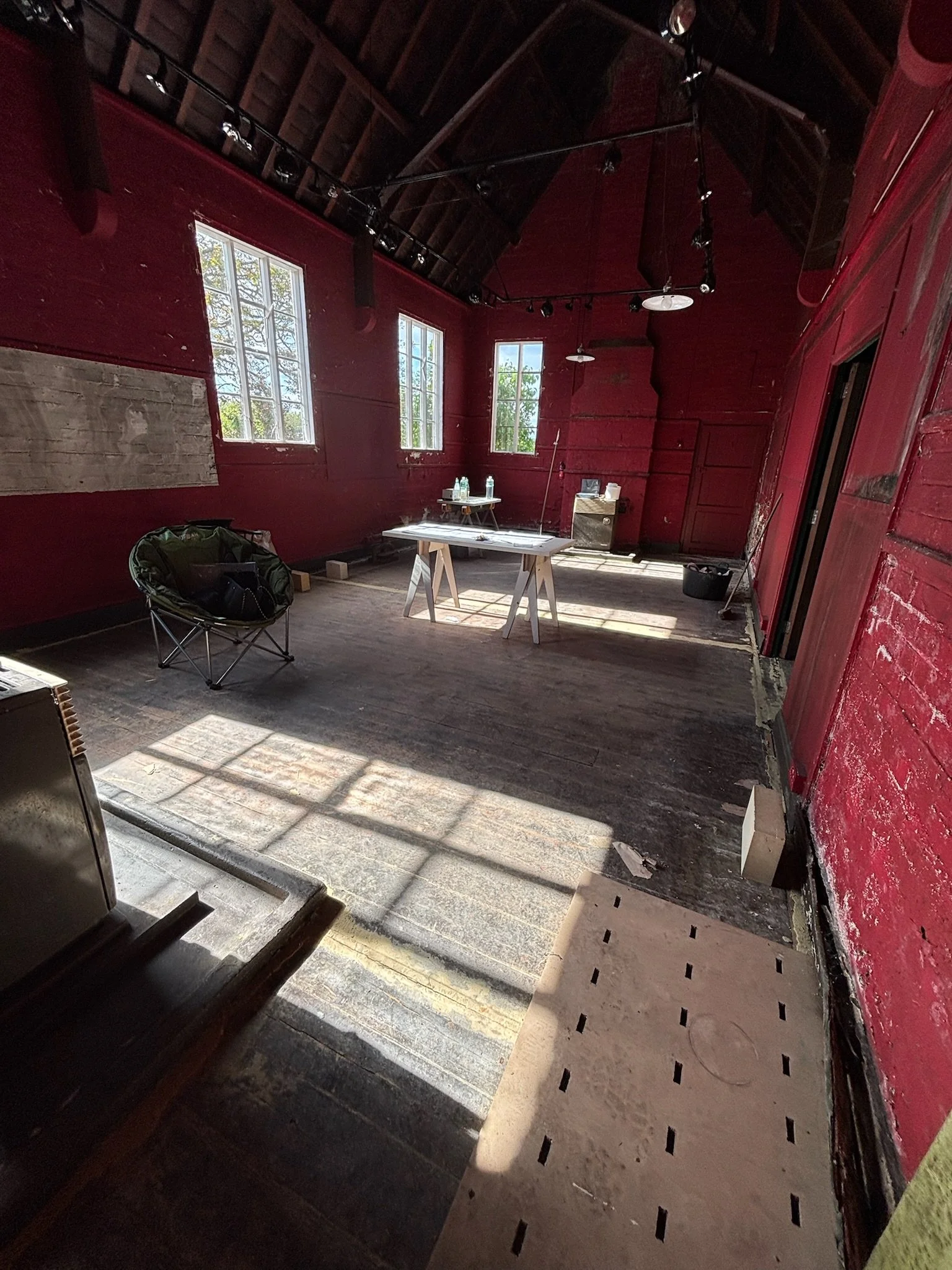 Empty room with red wooden walls, large windows letting in natural light, ceiling with exposed beams, and some furniture including a folding chair, a table, and a small cabinet.
