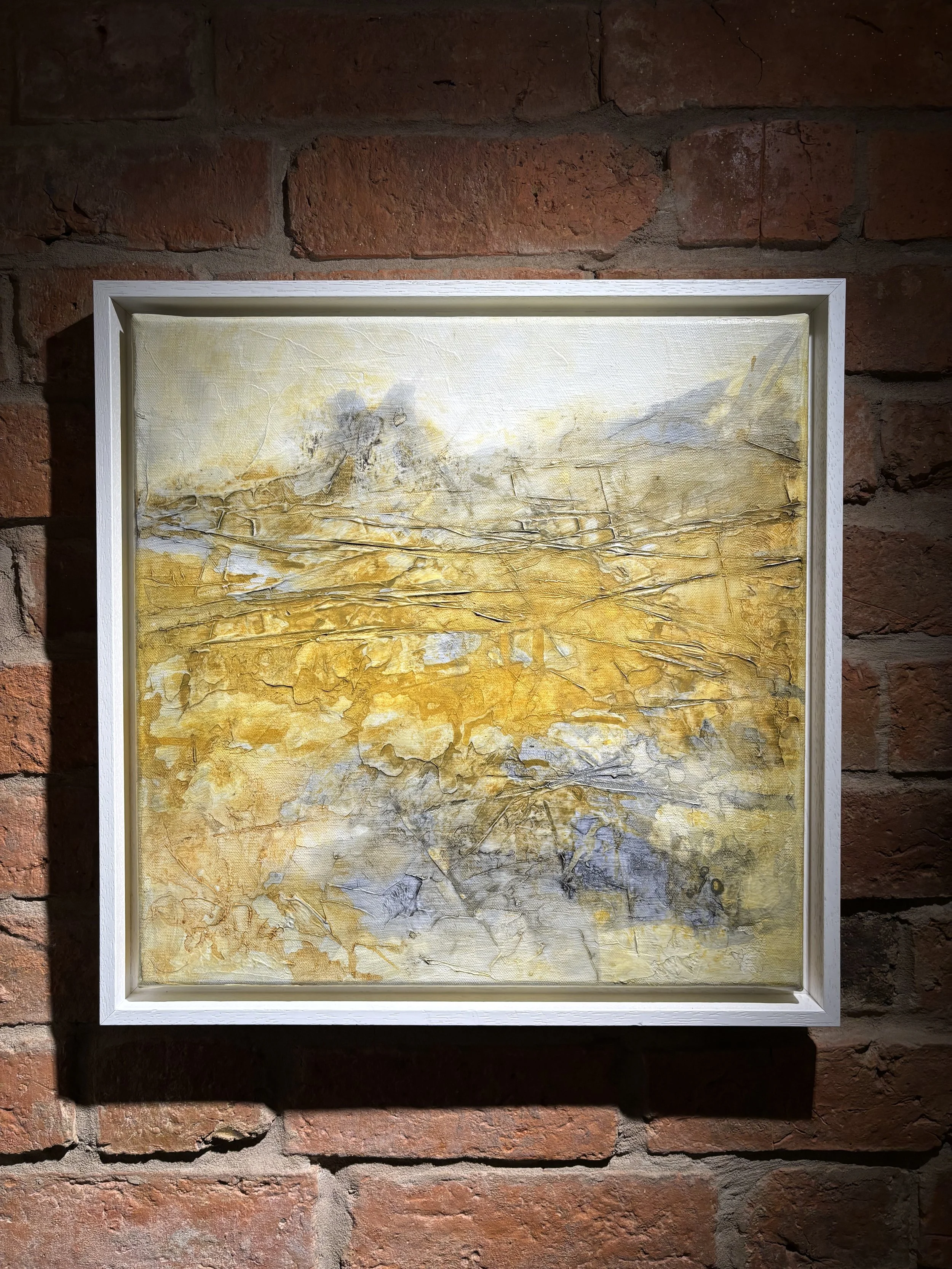Abstract landscape painting with earthy yellow, gray, and white tones, framed in white, hanging on a brick wall.