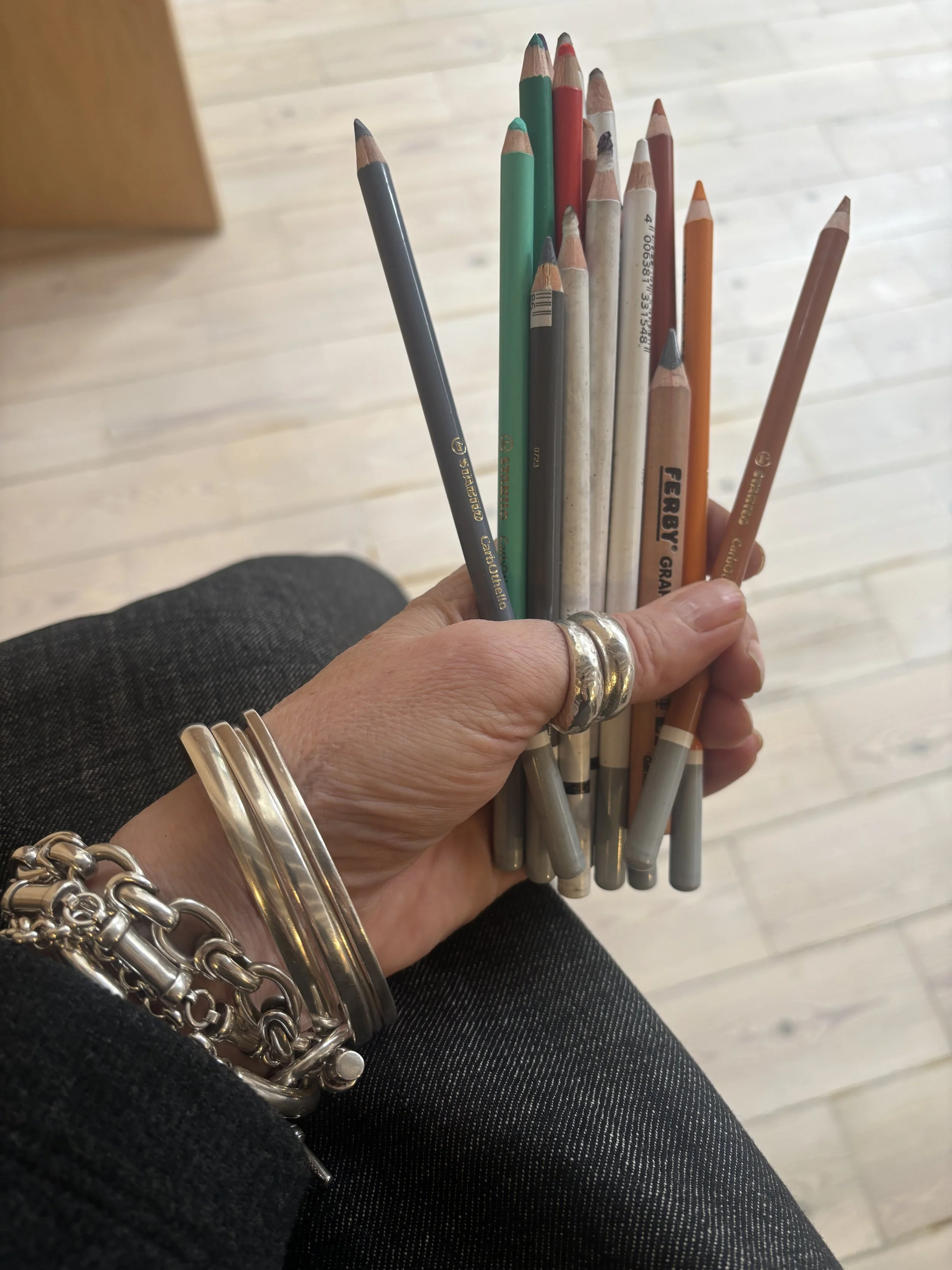 Hand holding a bunch of colored pencils with silver jewelry on wrist and fingers, background of light wood flooring.