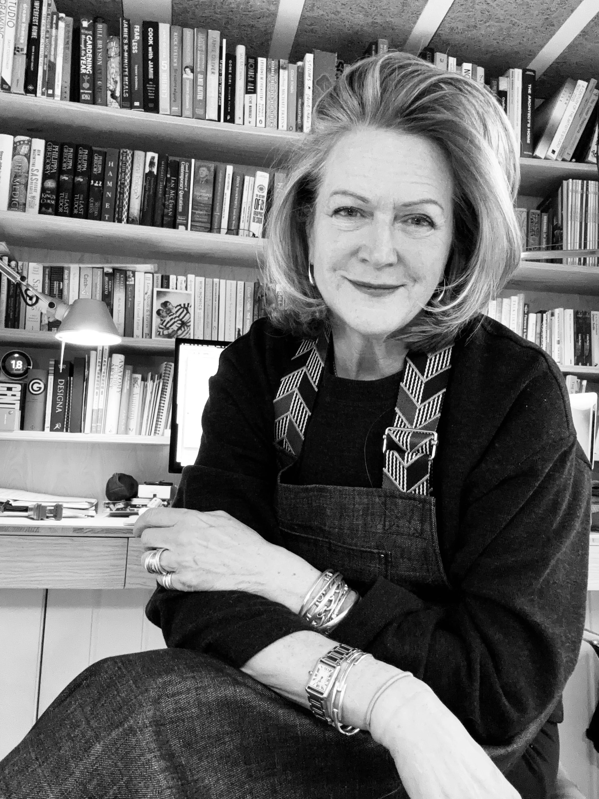 A woman with shoulder-length hair, wearing a dark top, sitting in front of a bookshelf filled with books, smiling at the camera.