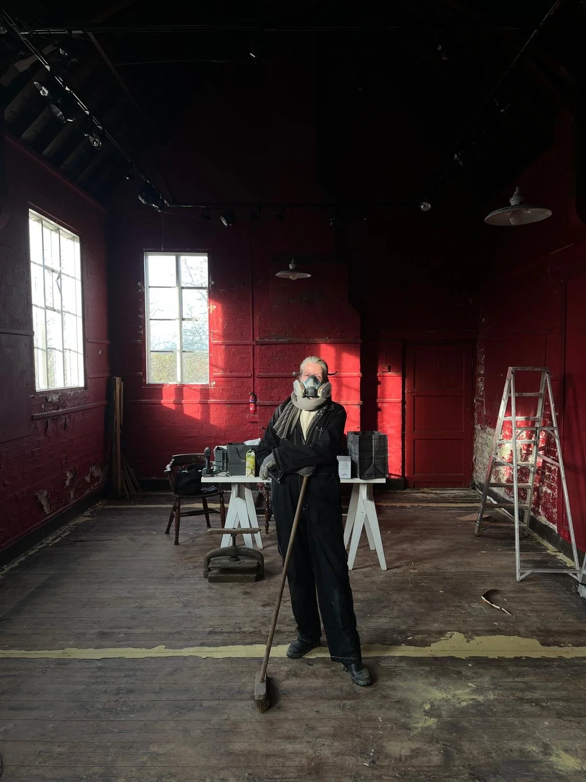 Person wearing a protective mask and dust mask standing in an empty room with red brick walls, holding a mop. The room has two windows letting in natural light, a stepladder, and various tools and supplies on a table.