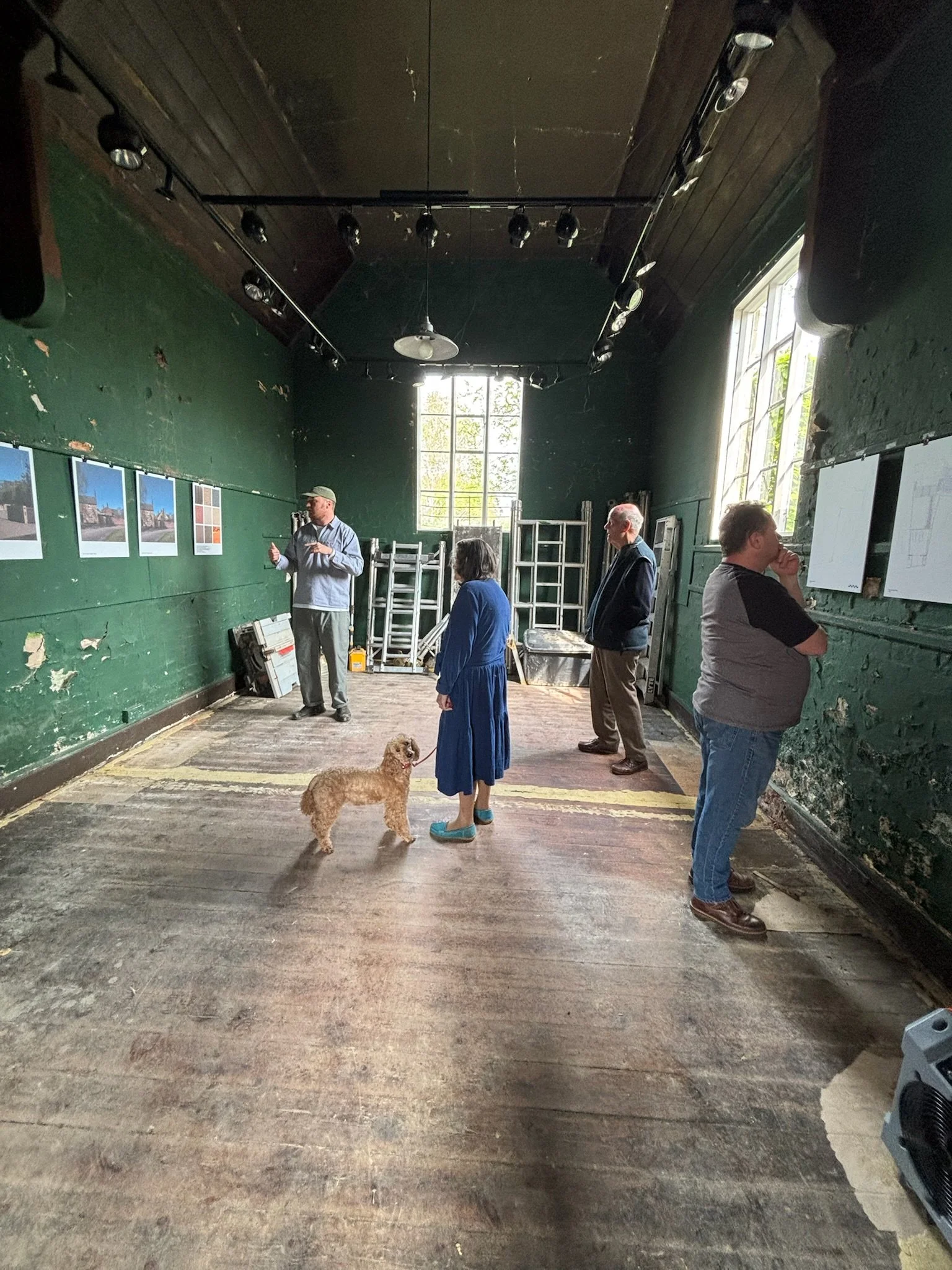 People in an art gallery with green walls, looking at photographs and sketches. One person is pointing, and a woman is standing with a dog in the center. The gallery has large windows, ceiling lights, and some scaffolding or ladders at the back.