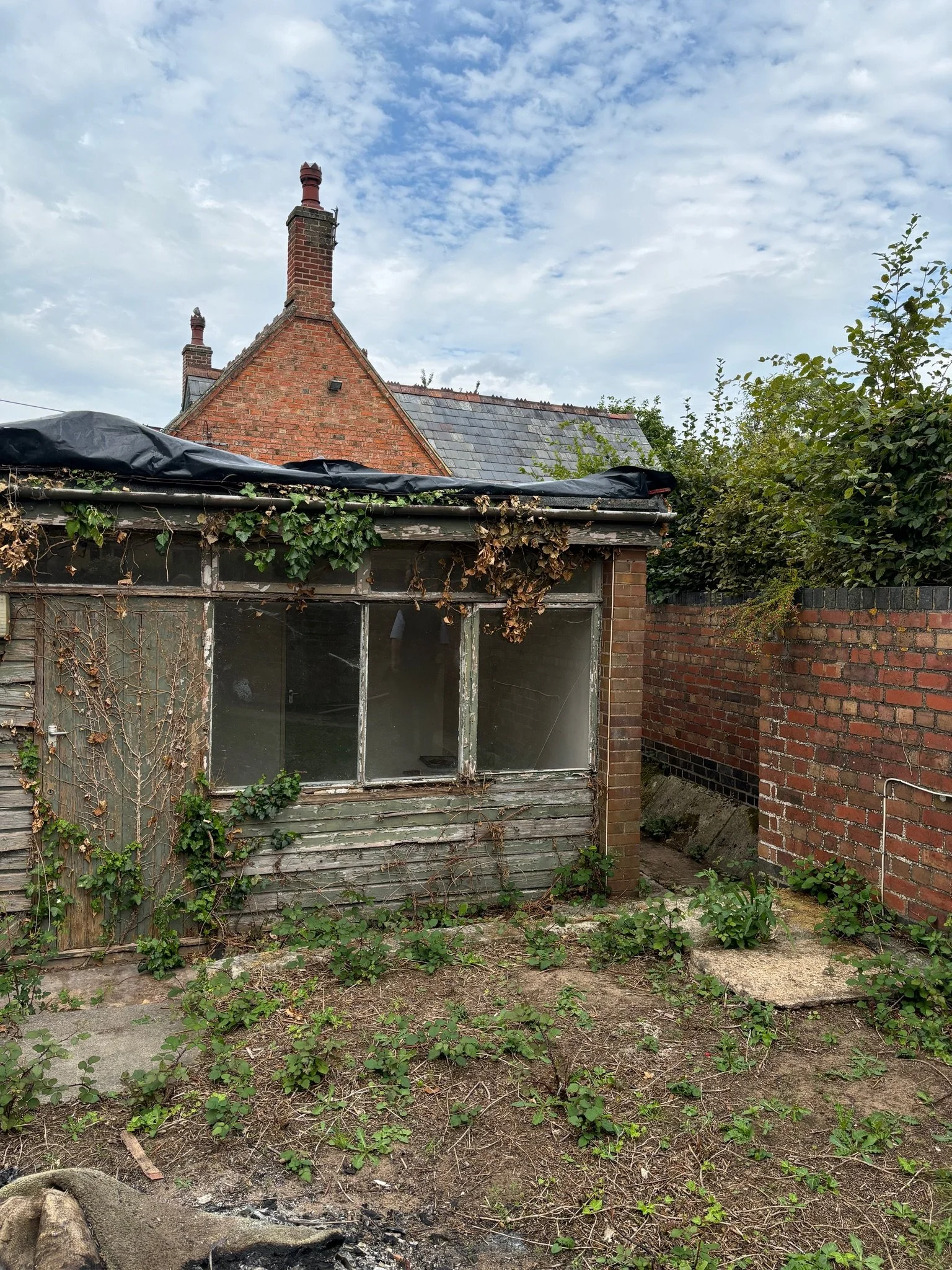 An old, weathered garden shed with peeling paint and overgrown plants, adjacent to a brick wall, with a cloudy sky overhead.