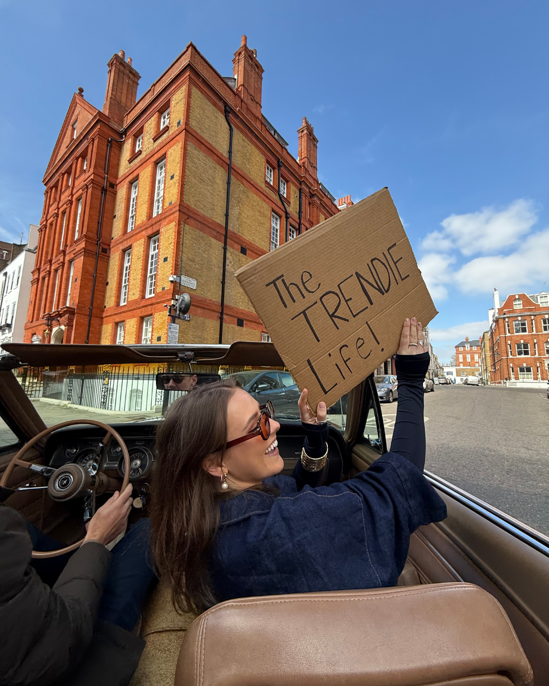 A woman with sunglasses and earrings in a vintage convertible car, holding a cardboard sign that says 'The TRENDIE Life!', smiling and looking to the side. The background shows a brick building and a clear blue sky.
