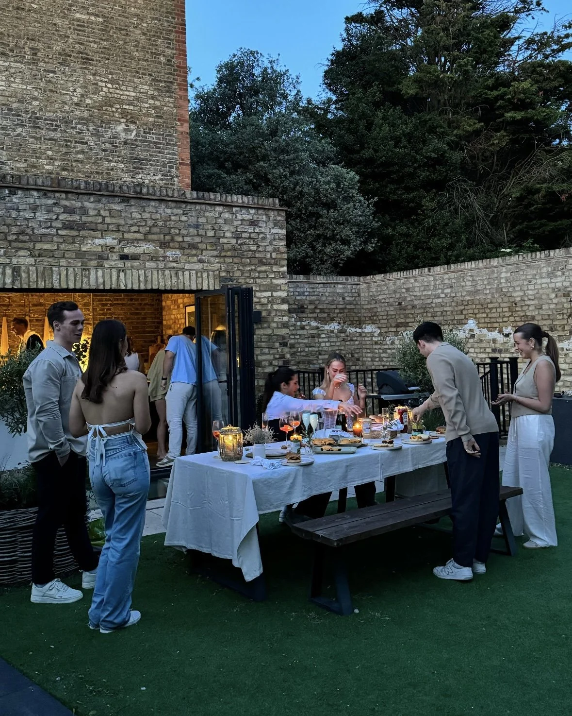 People enjoying a dinner party outdoors on a table with a white tablecloth, set with food, drinks, and candles, in a backyard with brick walls and trees at dusk.