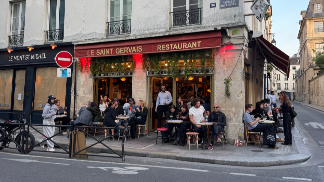 People dining outside at a restaurant on a city street in Paris, with signs reading 'Le Saint Gervais' and 'Le Mont St Michel.'