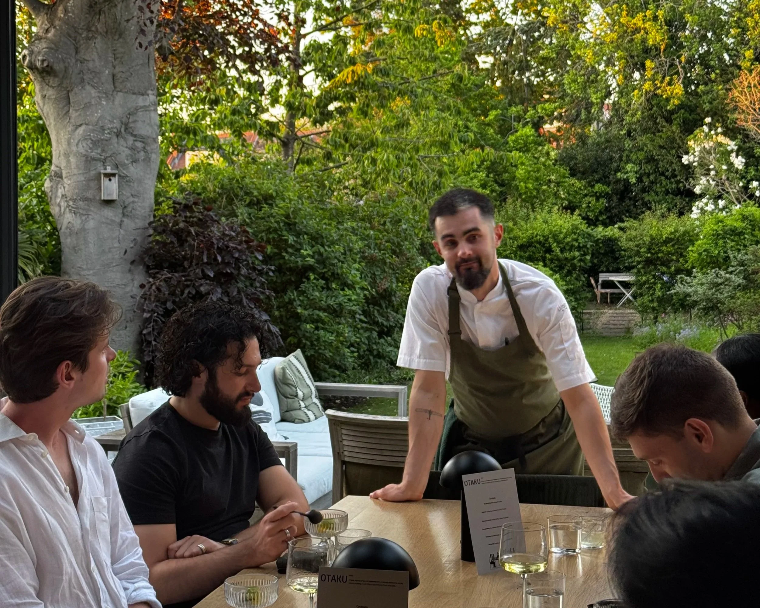 A group of five people, four men and one woman, sitting around a table outdoors in a garden setting. A man in a white shirt and green apron is standing and leaning forward with his hands on the table. The table is set with glasses of white wine, small bowls, and menus. The background features lush green trees and bushes, with open seating and a large tree to the left.