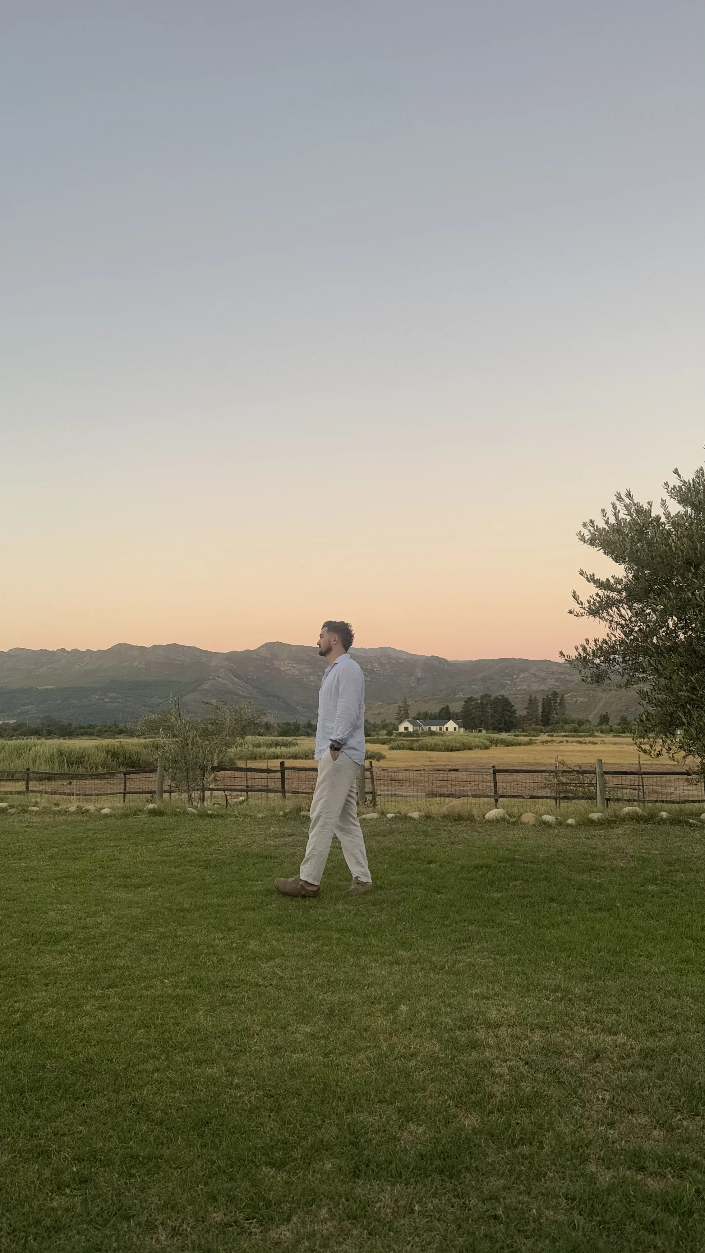 A man dressed in a white shirt and light-colored pants walking on green grass during sunset or sunrise, with mountains, trees, a fenced field, and a house in the background.