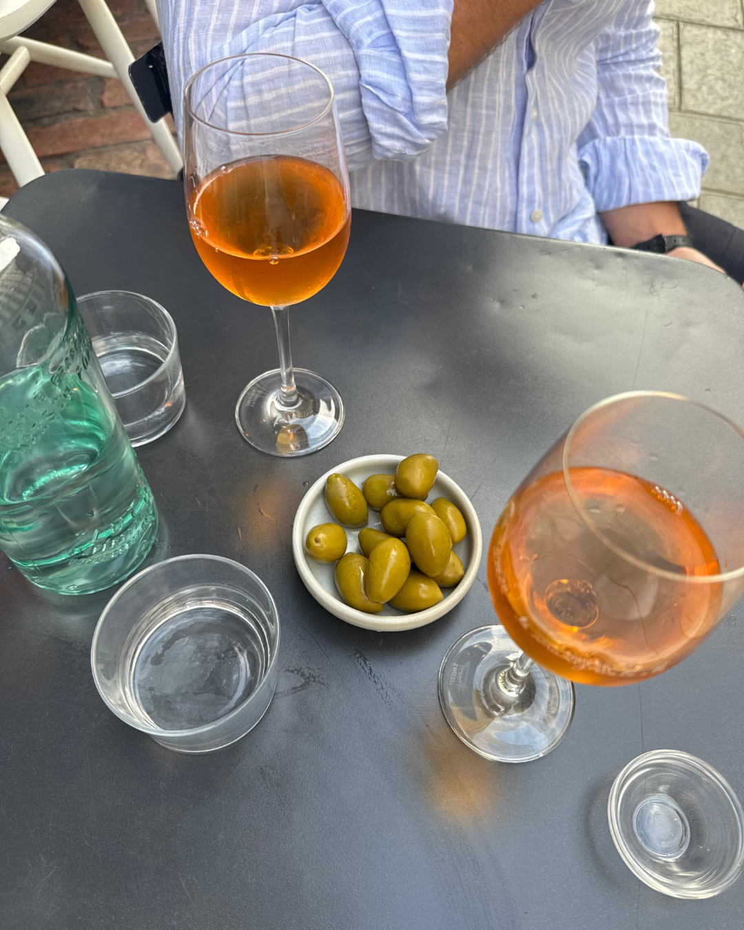 A table with two glasses of rosé wine, two glasses of water, a small bowl of green olives, a carafe of water, and an empty glass on a gray table. Part of a person's arm and torso in a blue striped shirt are visible in the background.