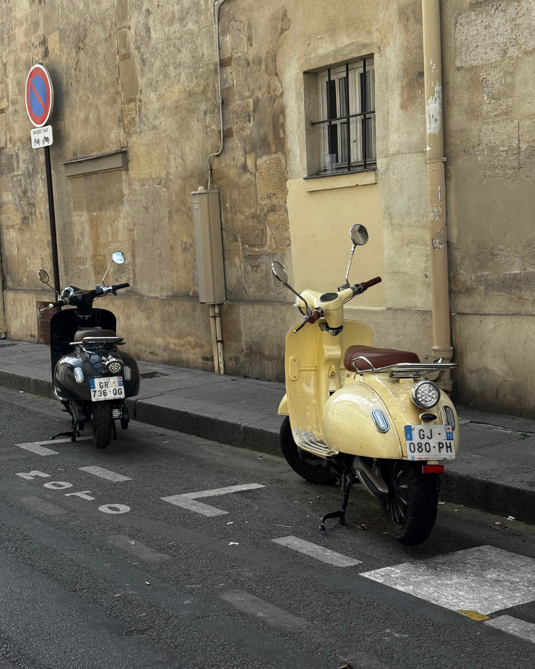Two vintage scooters parked on a street curb against an old stone building wall. One scooter is black with a rearview mirror and license plate, the other is yellow with a brown seat, mirror, and front light. A no-parking sign is mounted on a pole above them.