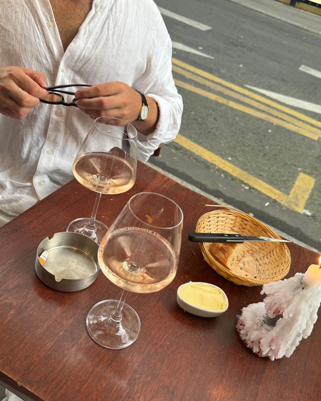 A person in a white shirt sitting at a wooden table outdoors with two glasses of rosé wine, a small dish of butter, a basket with bread, a candle, and an ashtray.
