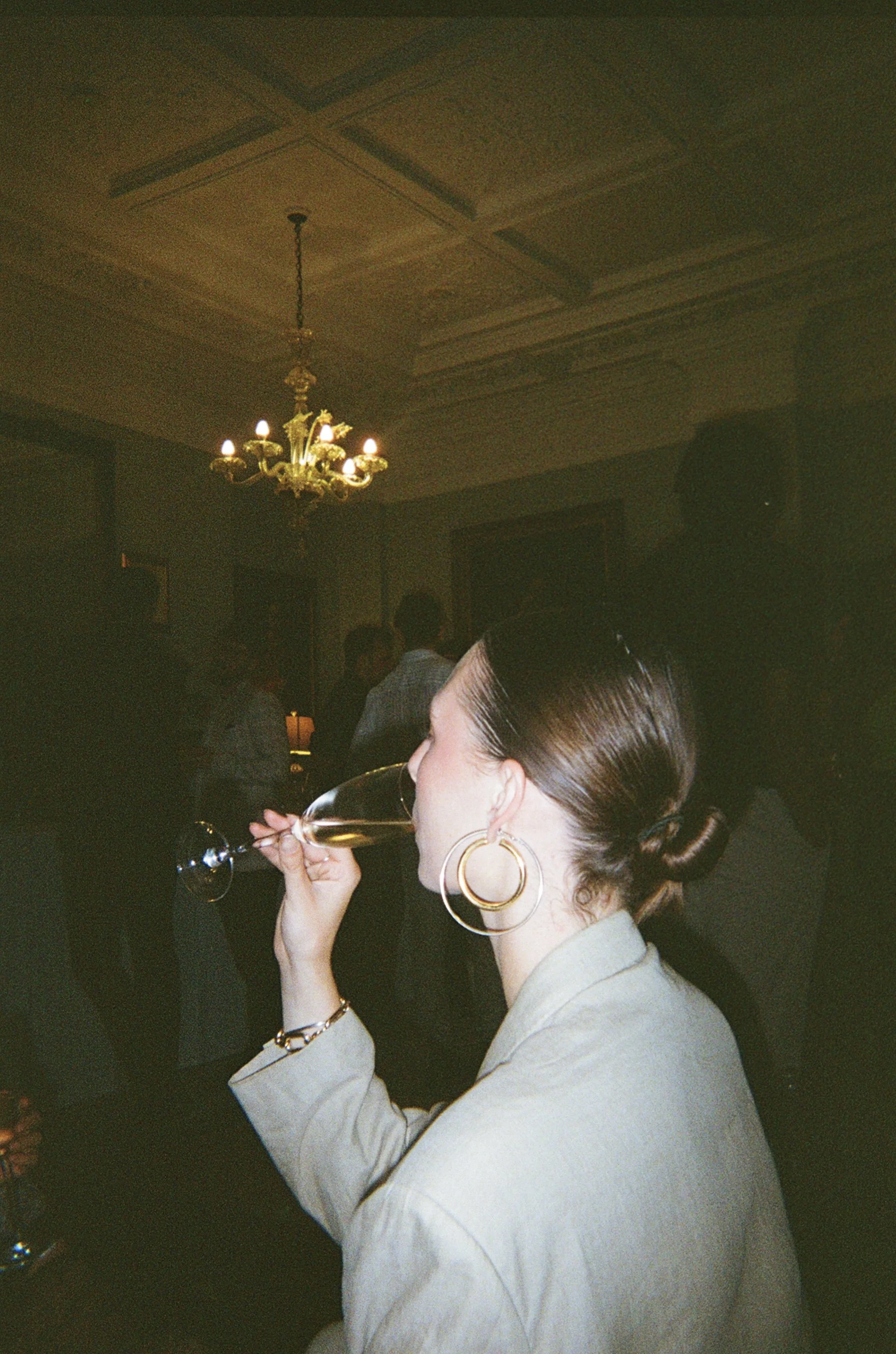 A woman with brown hair styled in an elegant bun wearing large gold hoop earrings and a beige blazer, holding a glass of white wine, at a dimly lit indoor gathering or party with people in the background.