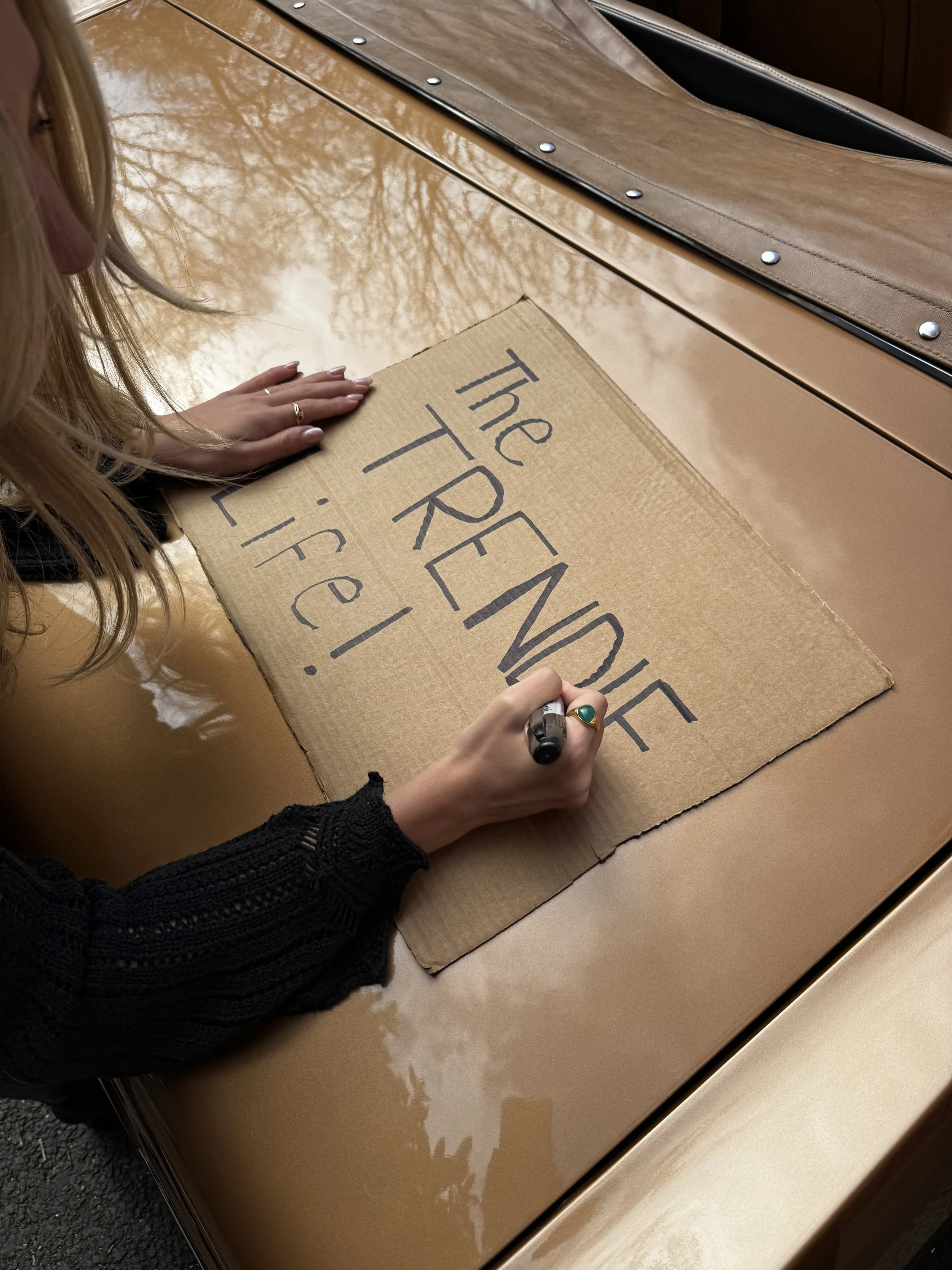 A person with long blonde hair and wearing a black knitted sweater is writing on a cardboard sign that says 'The FRIENDS TRAVEL Life!' with a black marker. The sign is placed on a shiny, tan-colored car hood, and the person's left hand is resting on the cardboard while their right hand writes.