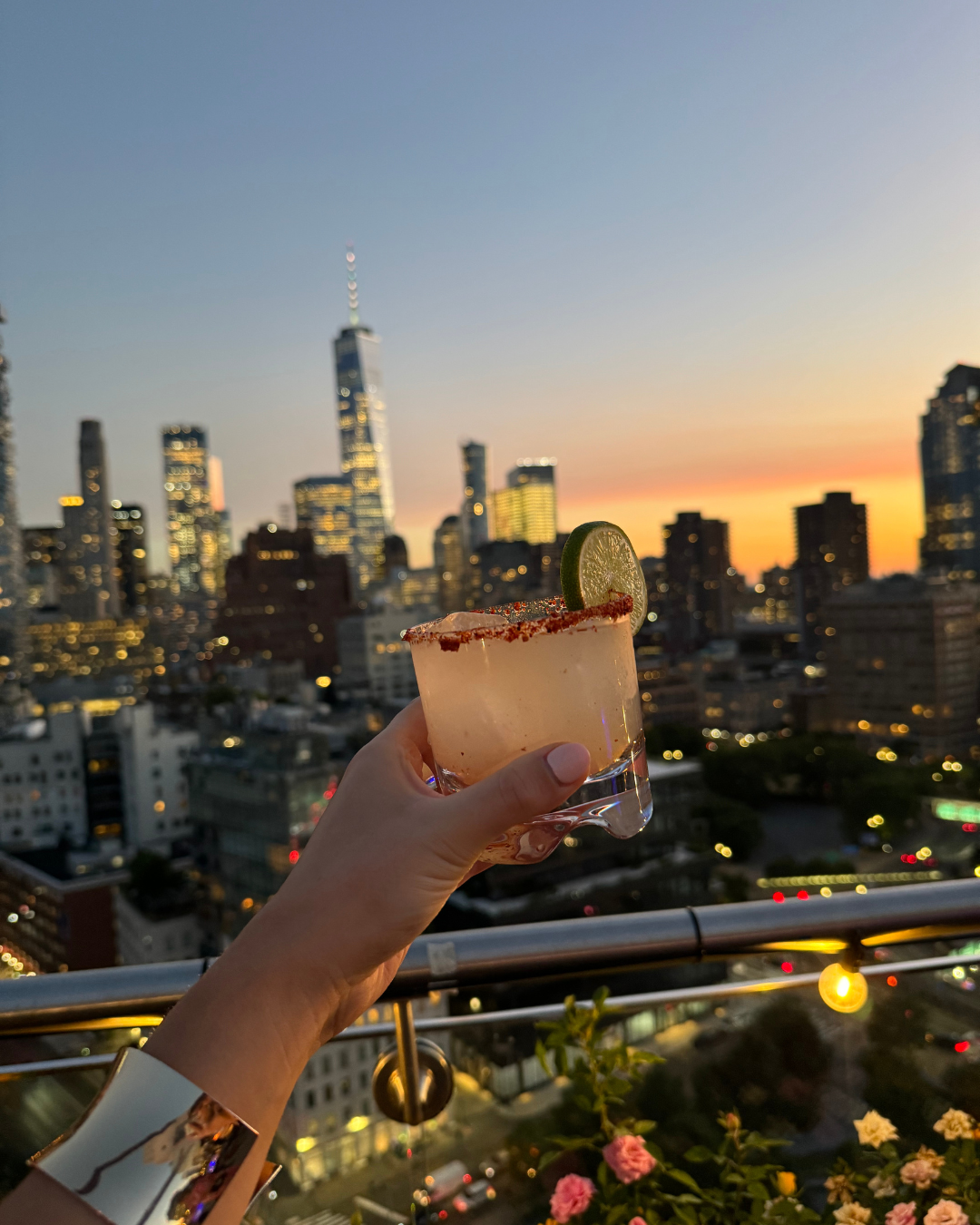 Person holding a margarita cocktail with a lime wedge against a city skyline during sunset.