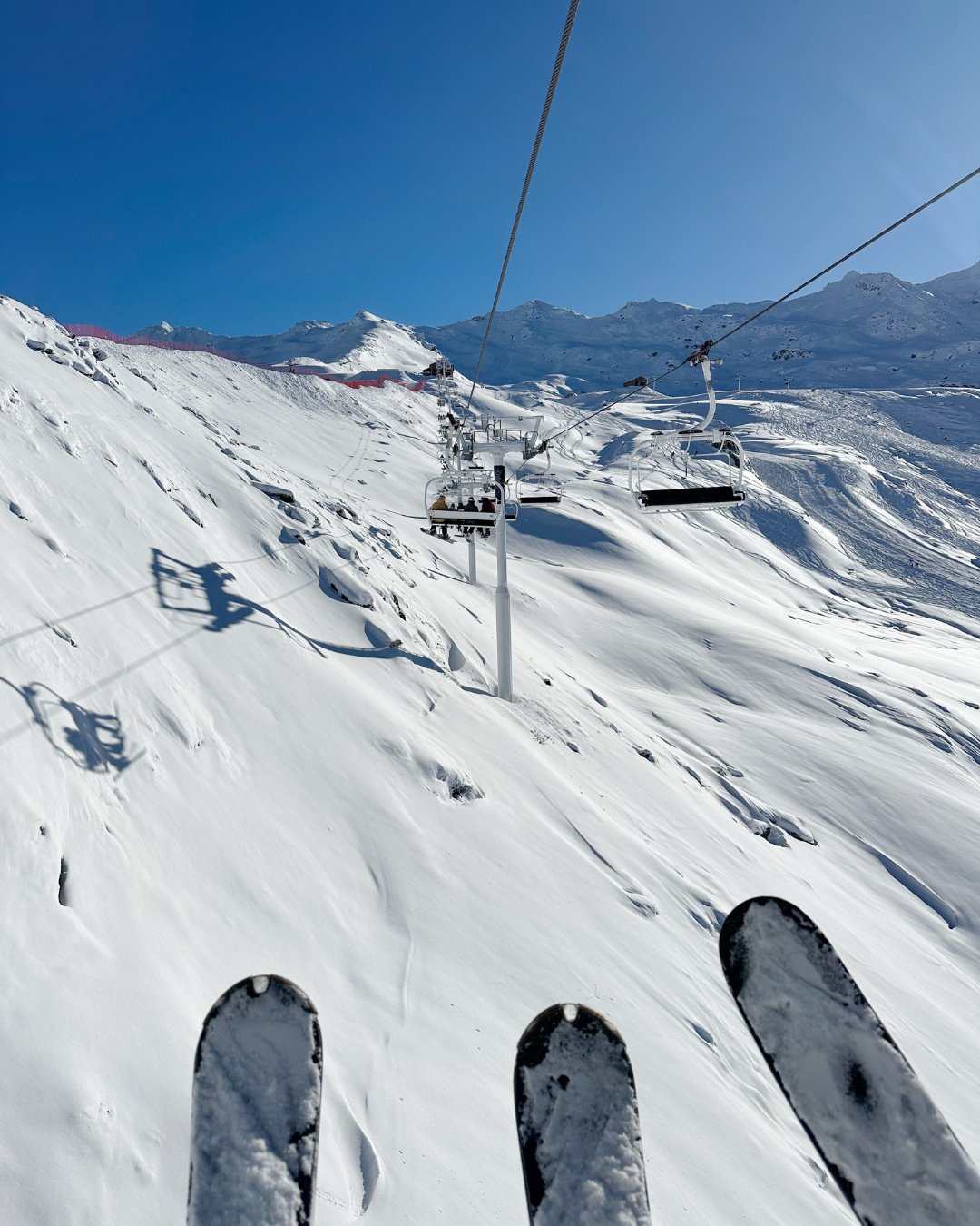 Skiers on a snowy mountain slope with ski lift chairs in the background, under a clear blue sky.