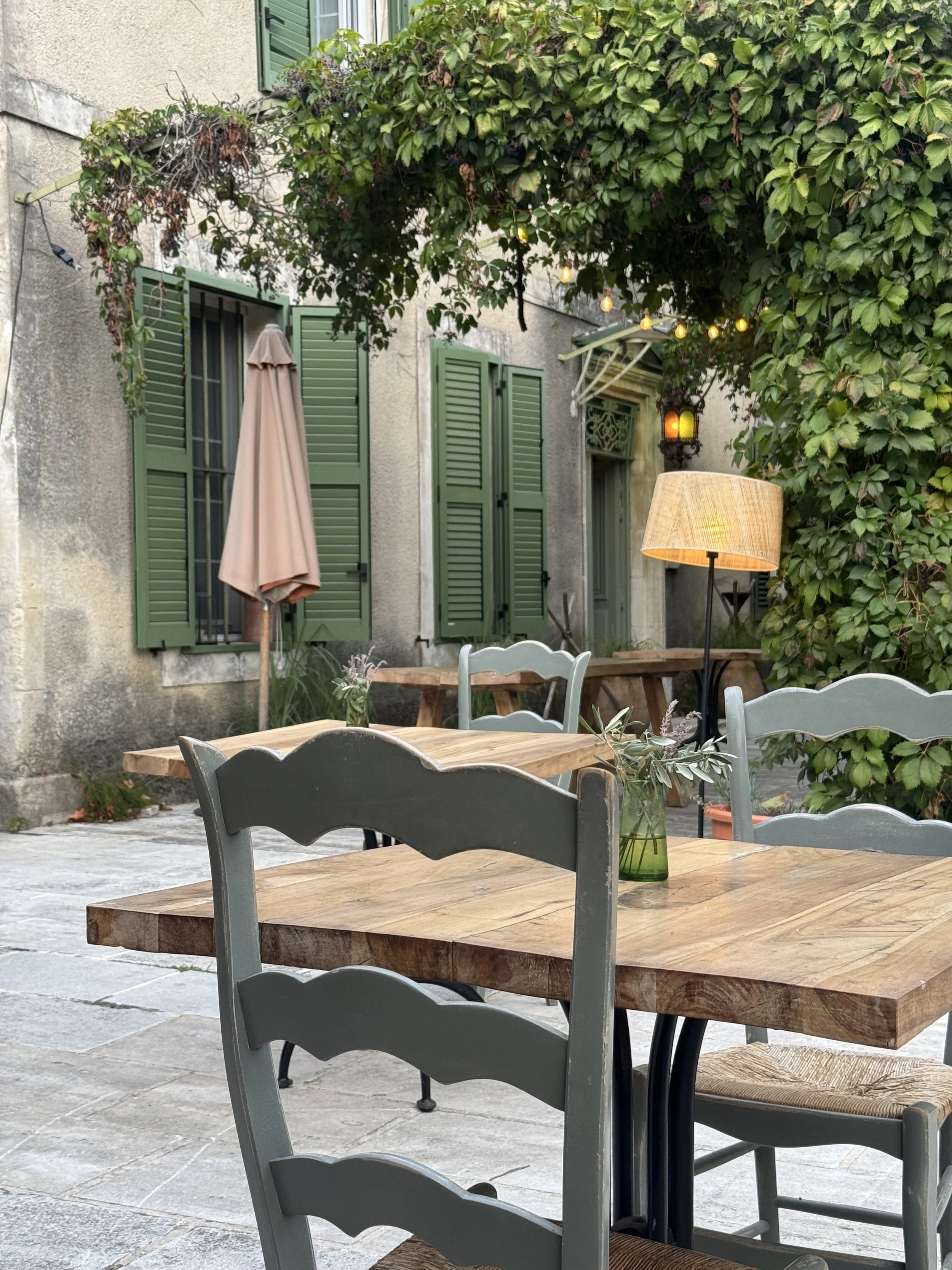 Outdoor patio with wooden tables, painted chairs, a lamp, a potted plant, string lights, a closed patio umbrella, green shutters on a stone building, and lush green foliage.