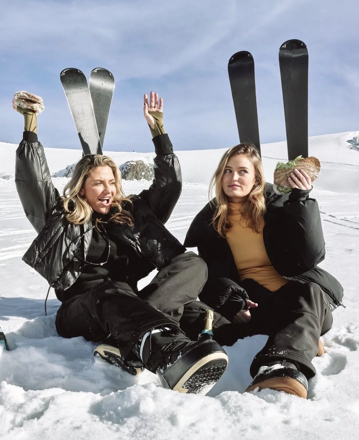Two women sitting in the snow in a winter landscape, holding skis and sandwiches, one making a playful gesture, both dressed in winter gear.