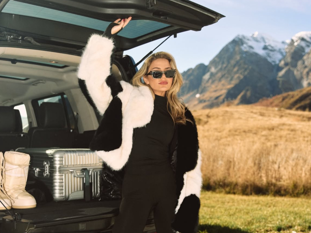 A woman in black clothing and sunglasses standing outdoors next to an open car trunk, with a coat hanging from her hand, and a mountainous landscape in the background.