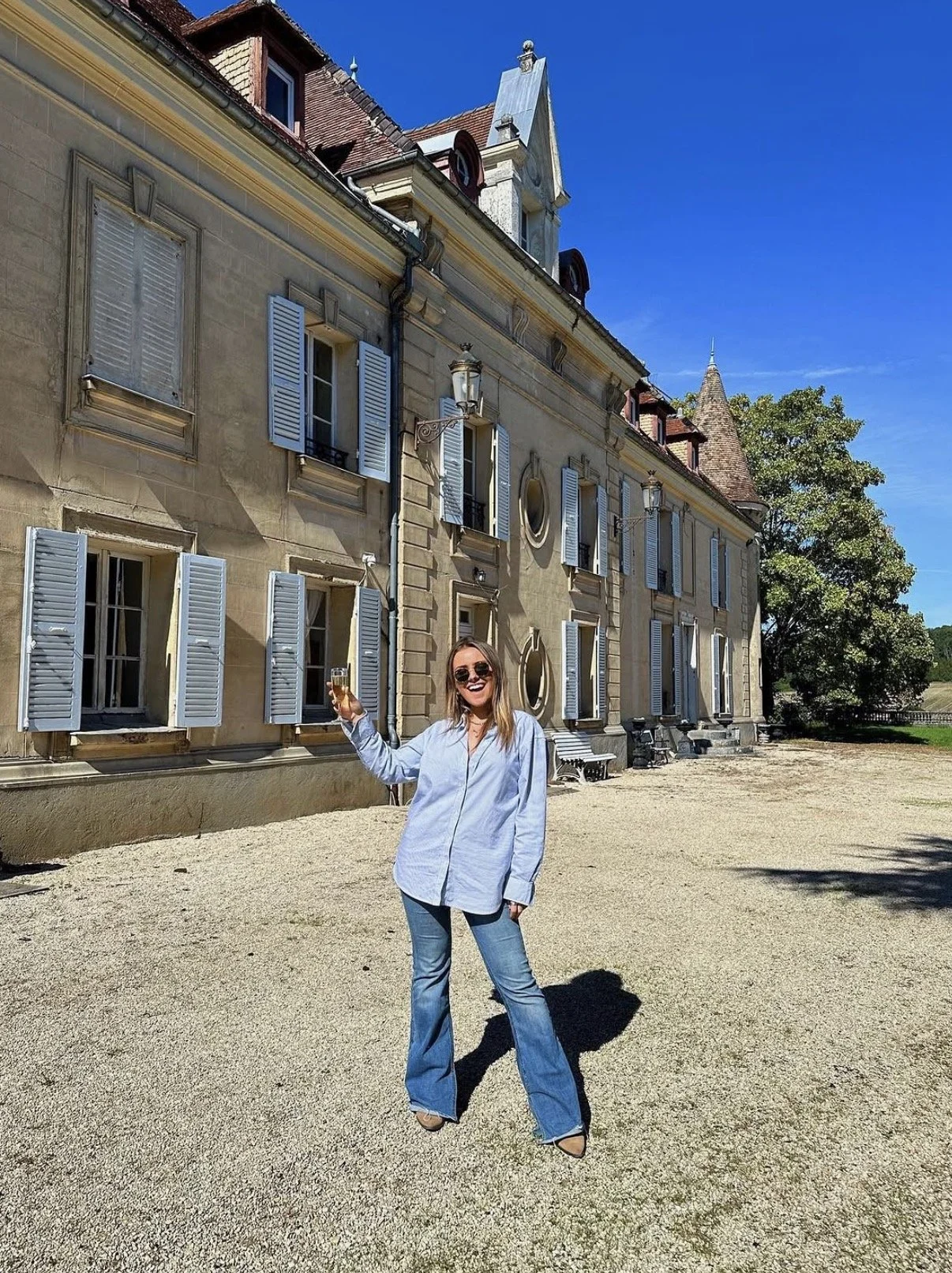 Woman with sunglasses, smiling, wearing a light blue shirt and jeans, holding a glass of champagne outdoors in front of a historic European building with beige walls and white shutters, under a clear blue sky.