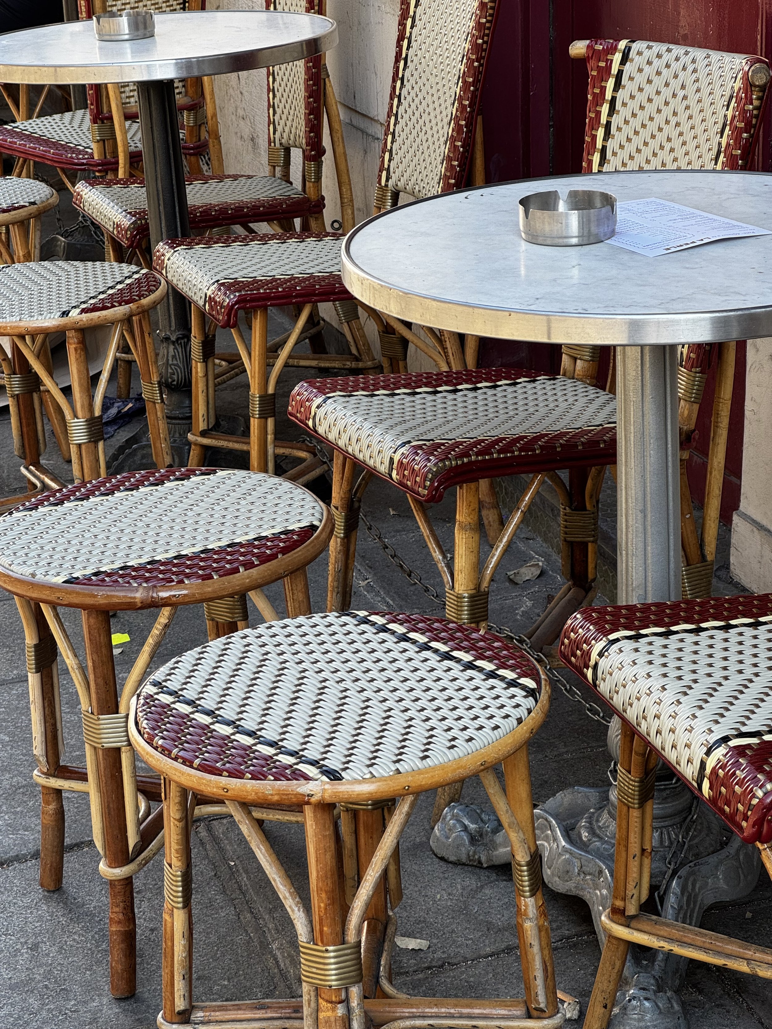 Empty outdoor cafe tables with wicker chairs, one with a metal ashtray and a menu, set on a sidewalk outside.