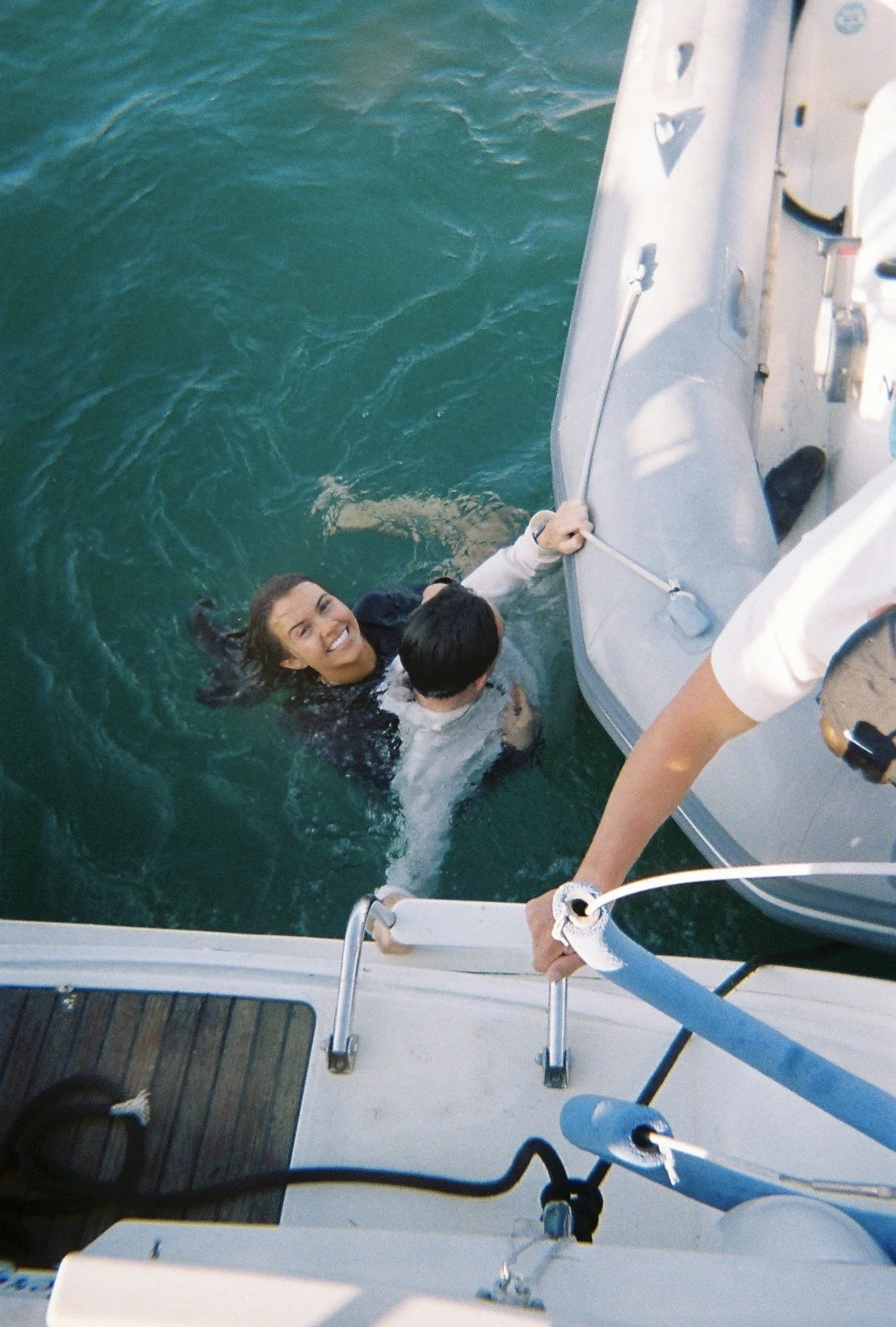 A woman smiling in the water holding onto the side of a boat, with a person next to her, while another person on the boat reaches down to assist.