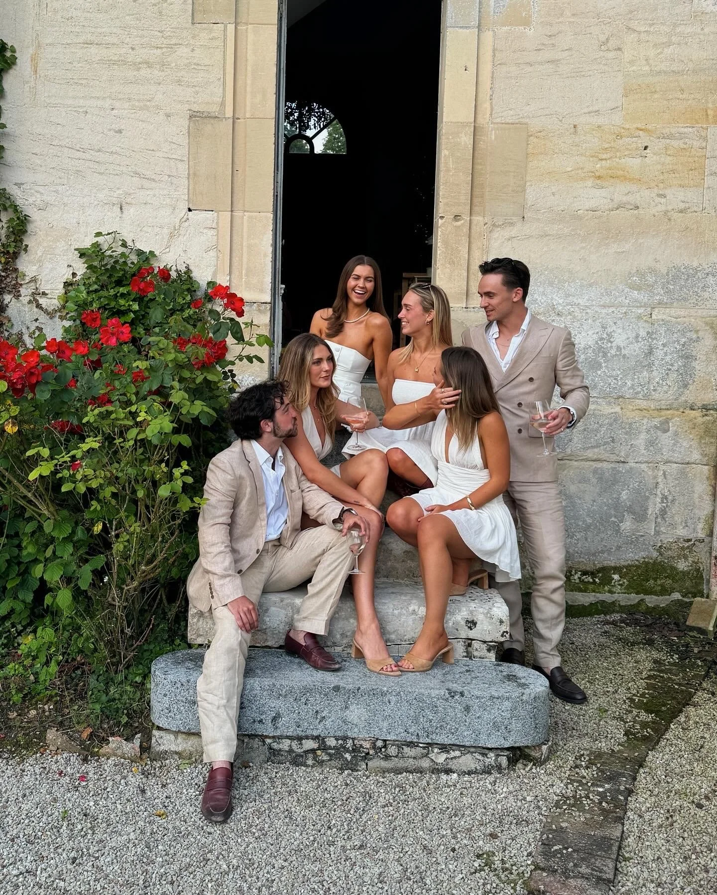 A group of six young adults in formal attire gathered outside a historic stone building, with three women and two men holding glasses of wine, engaged in conversation, and one woman seated on a stone bench laughing.