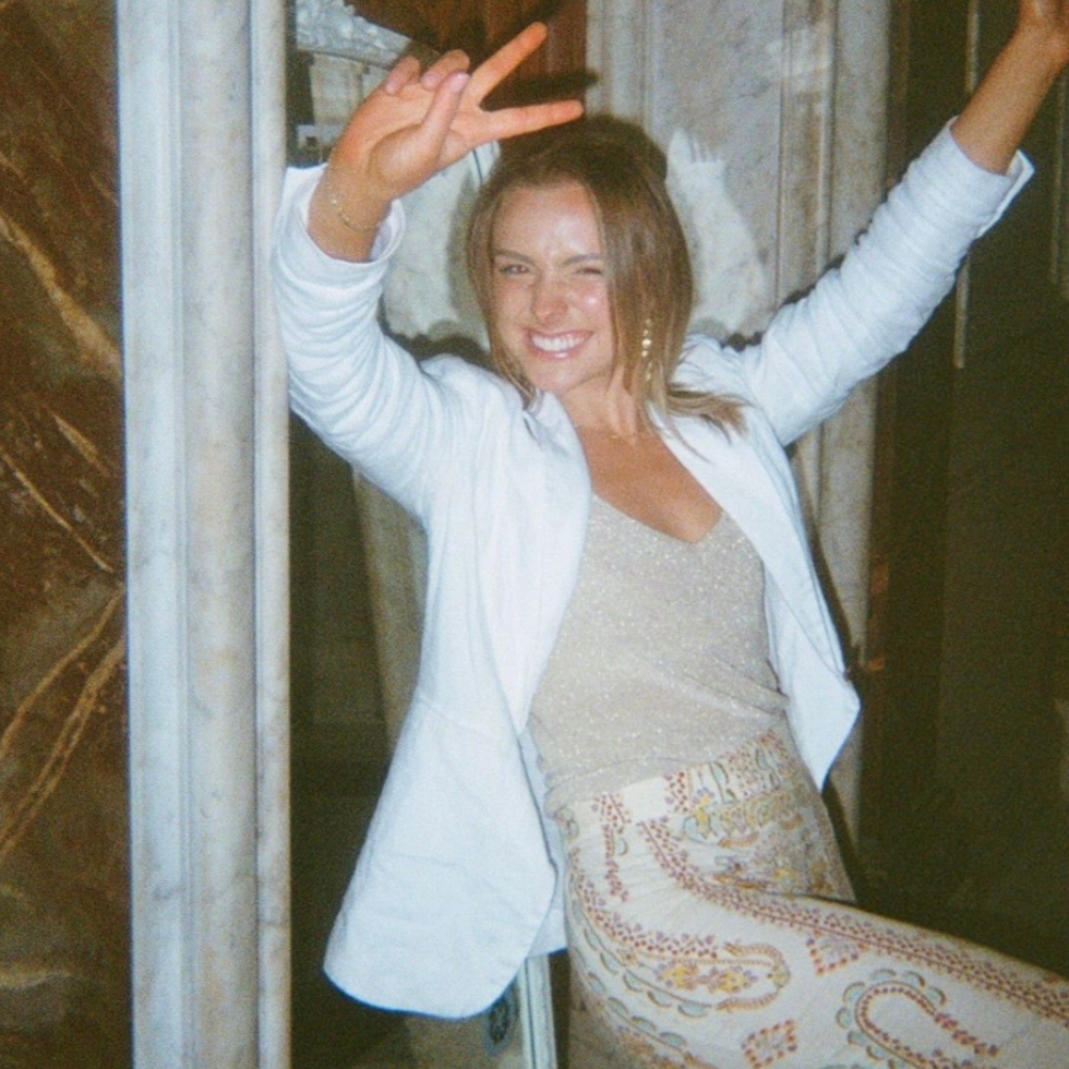 A woman smiling and making a peace sign with her right hand, wearing a white cardigan over a beige top and patterned pants, standing indoors next to a window with blinds.