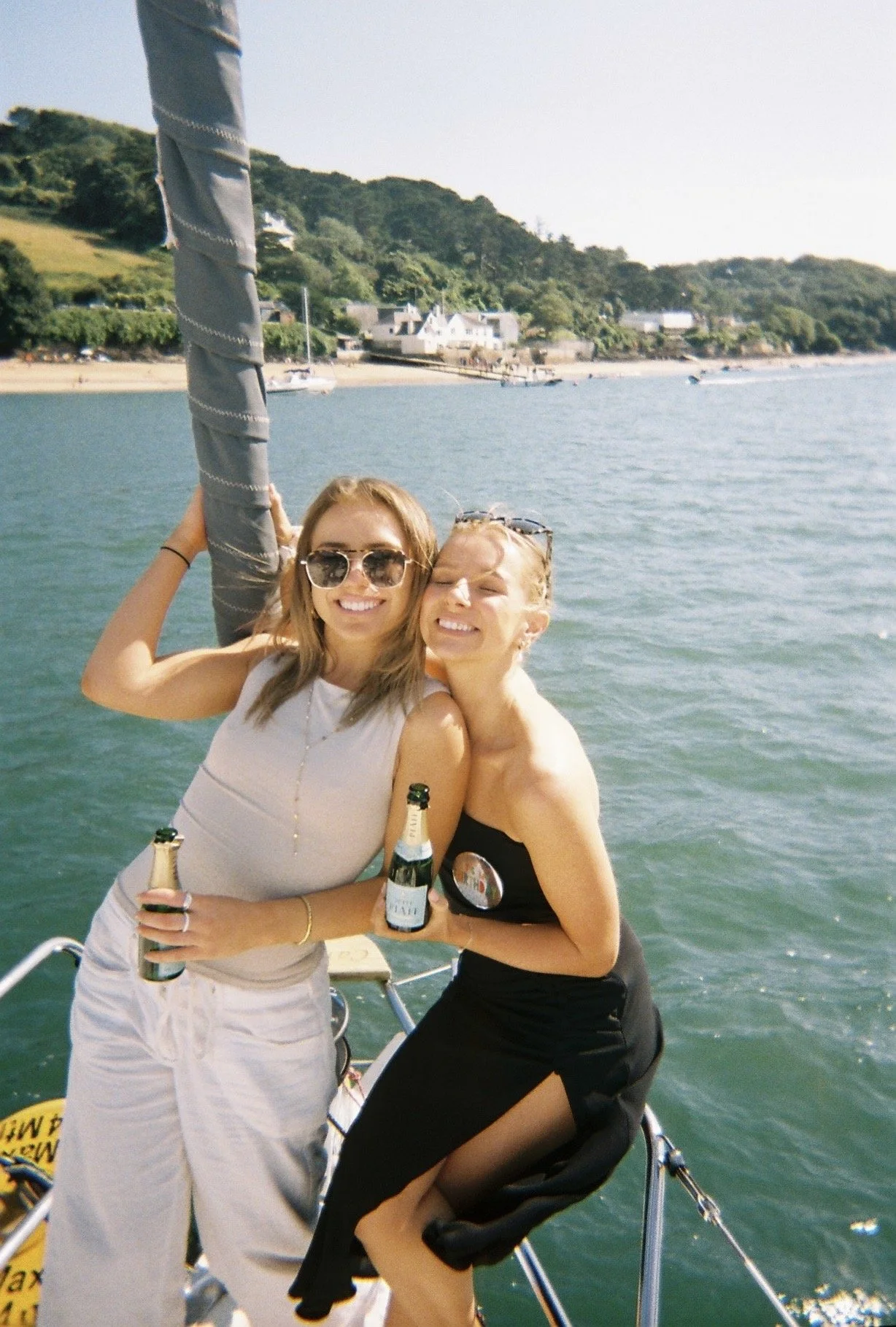 Two young women smiling on a boat, holding bottles of champagne, with a scenic water and shoreline background.