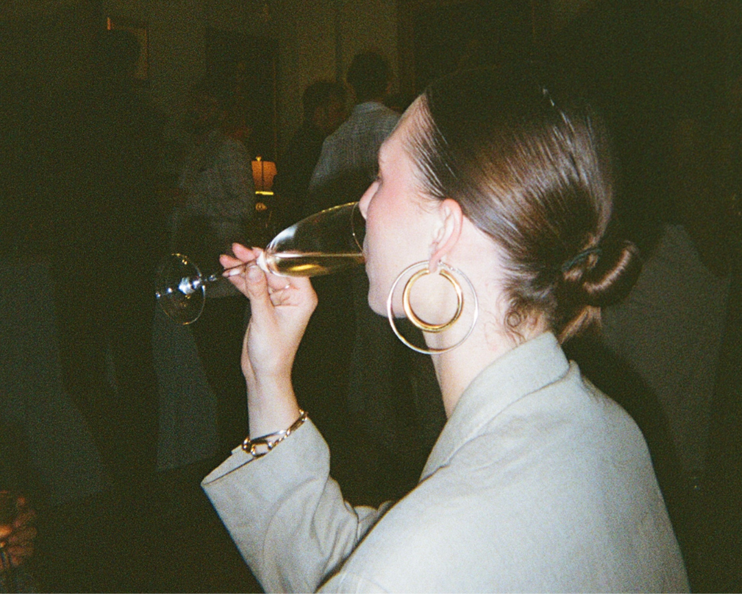 A woman with dark hair in a bun wearing large gold hoop earrings and a gray blazer, drinking champagne from a flute glass at a social event or party.