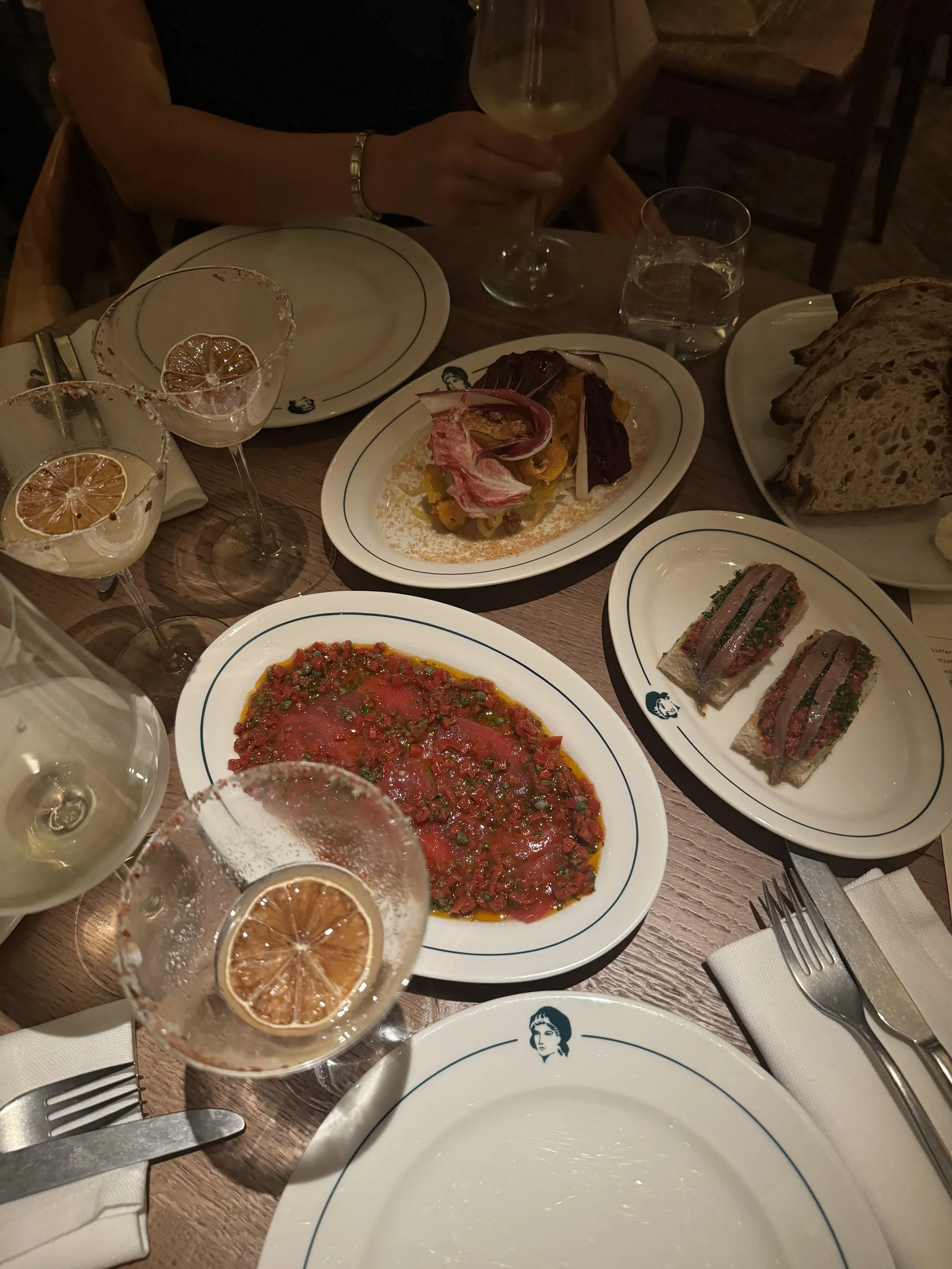 A dinner table set with multiple plates of food, including a sliced loaf of bread, a tomato-based appetizer, a small open-faced sandwich with meat and greens, and a colorful vegetable dish. There are also glasses of white wine, a glass of water, and cocktail glasses with lemon slices. A person is visible holding a wine glass.