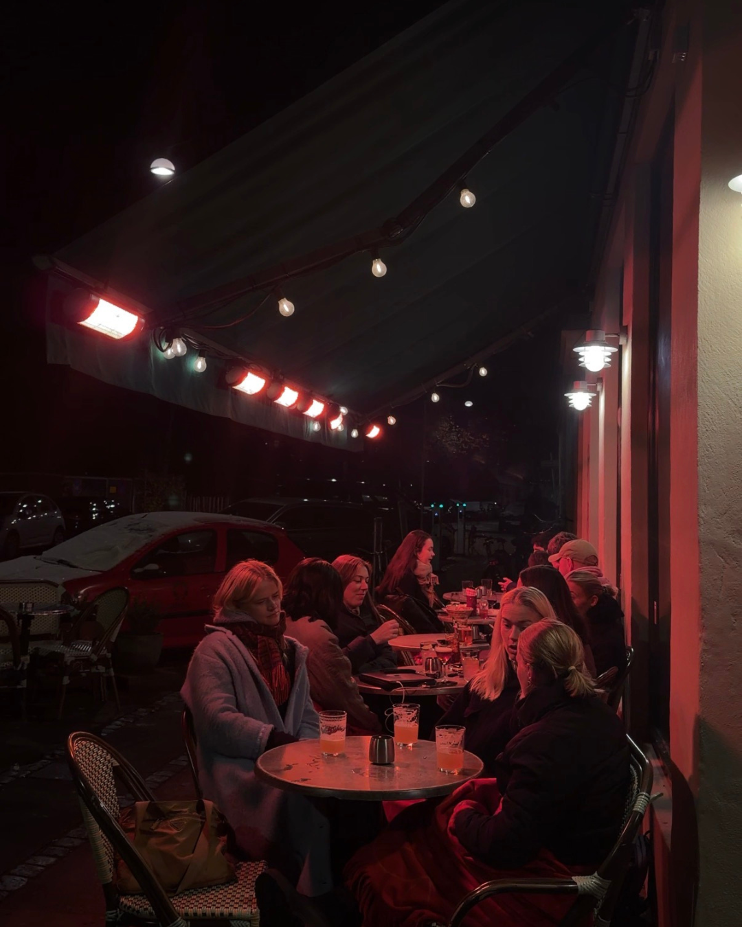 People sitting at tables outside a restaurant at night, under red and white string lights and wall-mounted lamps, with parked cars in the background.