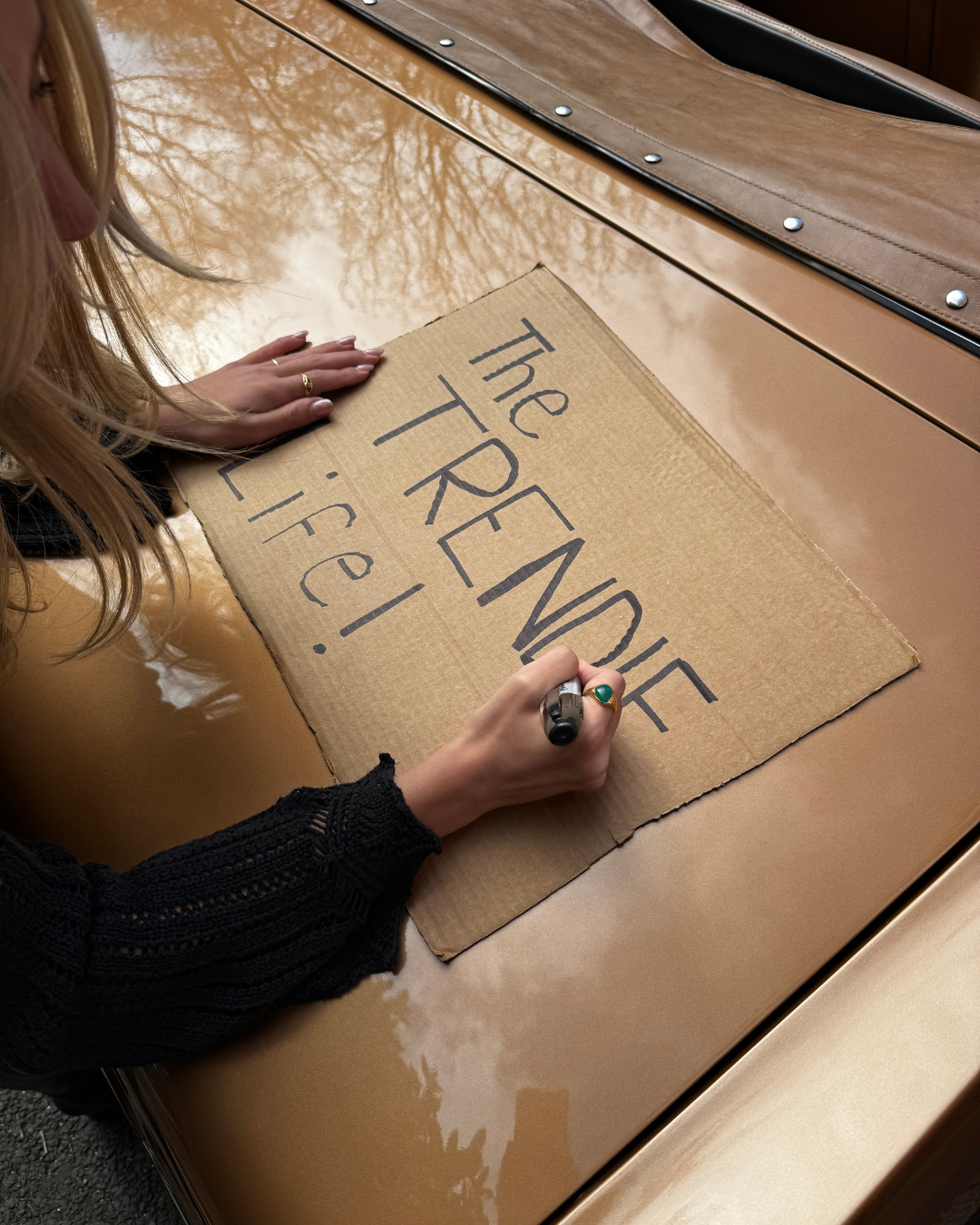 A person with blonde hair, wearing a black sleeve with lace, writes on a cardboard sign with a black marker that says, 'The REDEFINE TRUTH Life!' The person is sitting at a tan surface, possibly a car, with a reflection of tree branches visible.