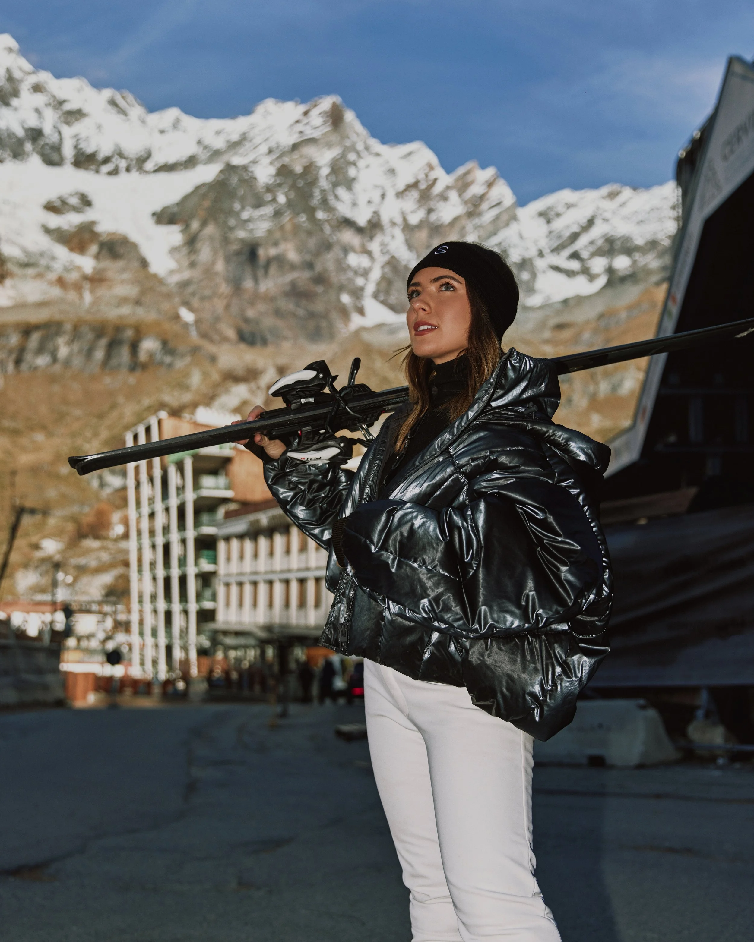 A woman in a black jacket, white pants, and black beanie, holding a ski pole, standing outdoors with snowy mountains in the background.