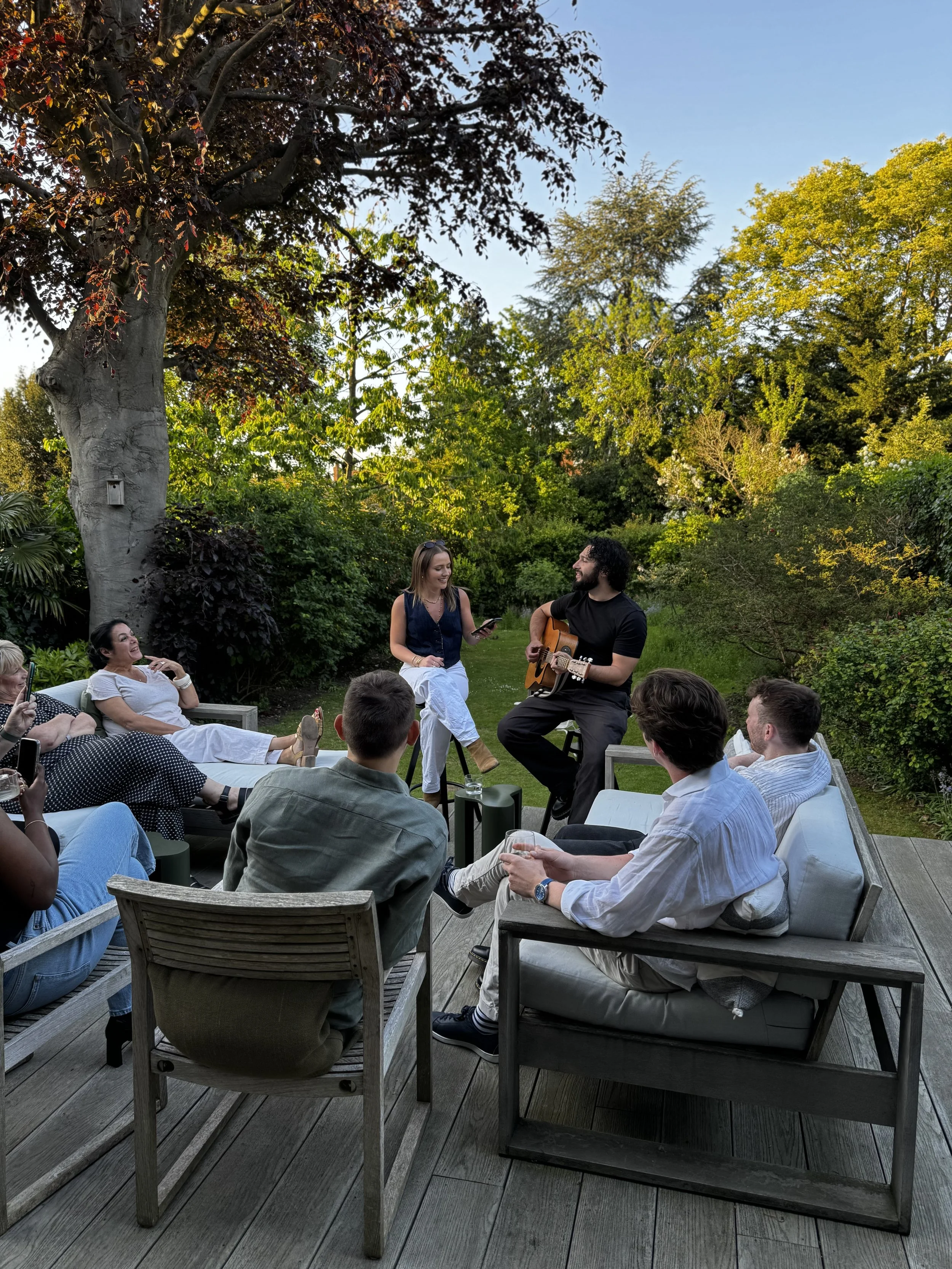 People enjoying an outdoor gathering in a lush, green garden during the evening. A man is playing guitar, while a woman is singing or speaking into a microphone. Others are sitting on outdoor furniture, watching and listening. There is a large tree and various shrubs in the background.