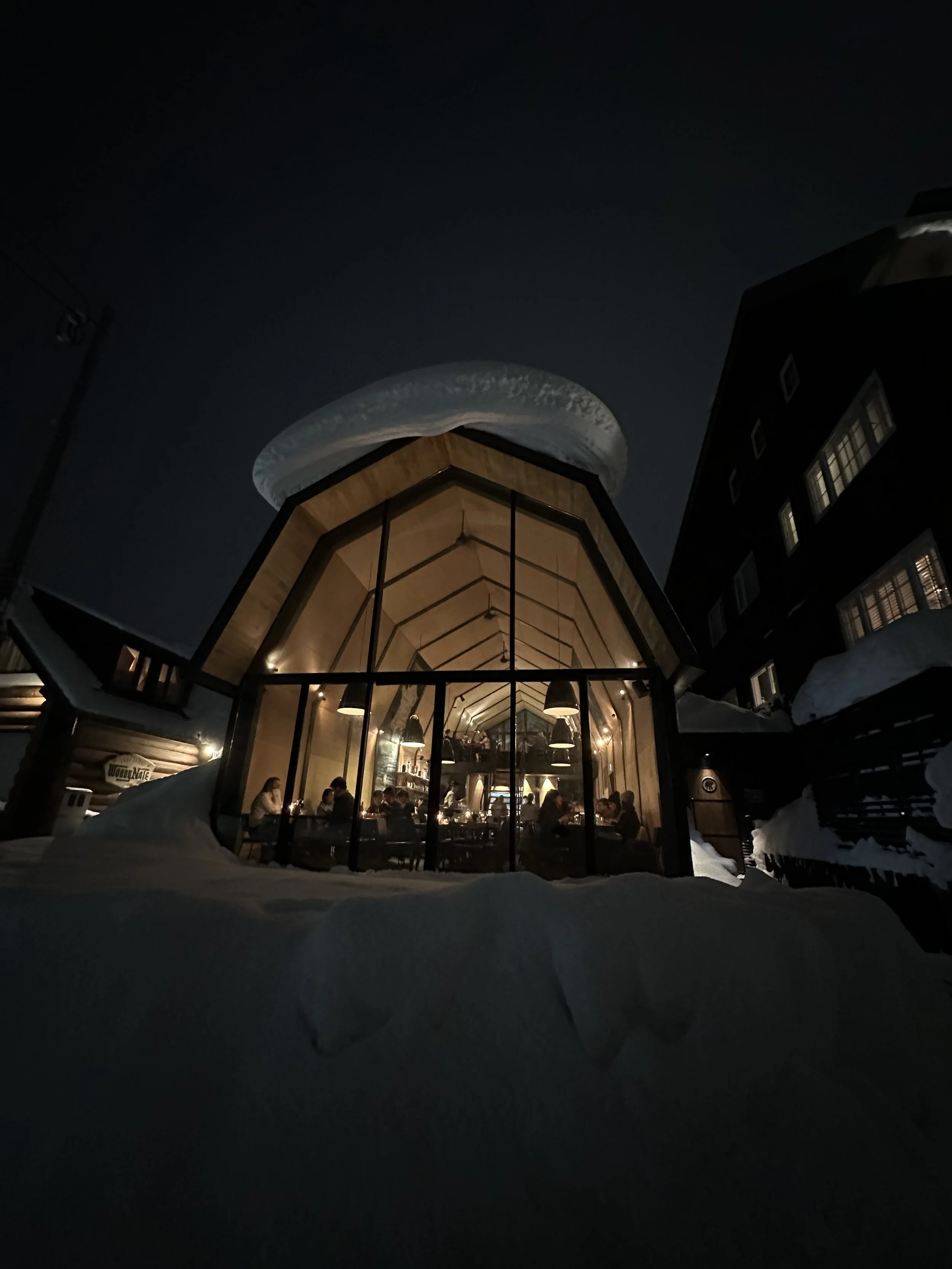 Nighttime view of a glass-walled restaurant or cafe in a snowy outdoor setting, with warm lighting and people dining inside, surrounded by snow-covered ground and nearby buildings.