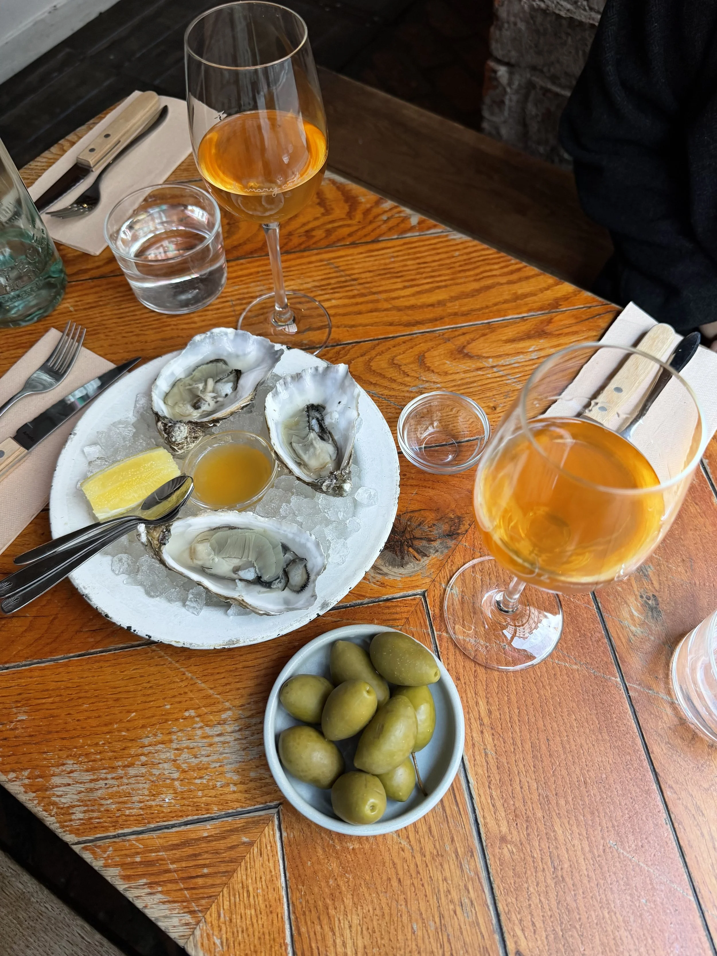 A table with a seafood appetizer of oysters on a bed of ice, lemon wedge, and small bowl of sauce, green olives in a separate bowl, two glasses of rosé wine, a glass of water, utensils, and napkins.