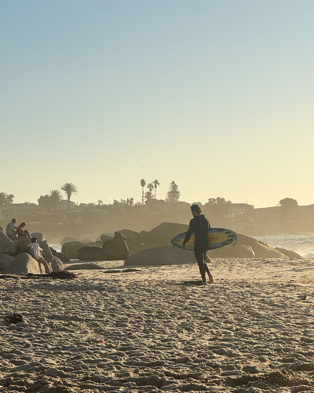 A person walking on the beach with a surfboard, back to camera, during sunset or sunrise, with other people sitting on rocks and palm trees in the background.