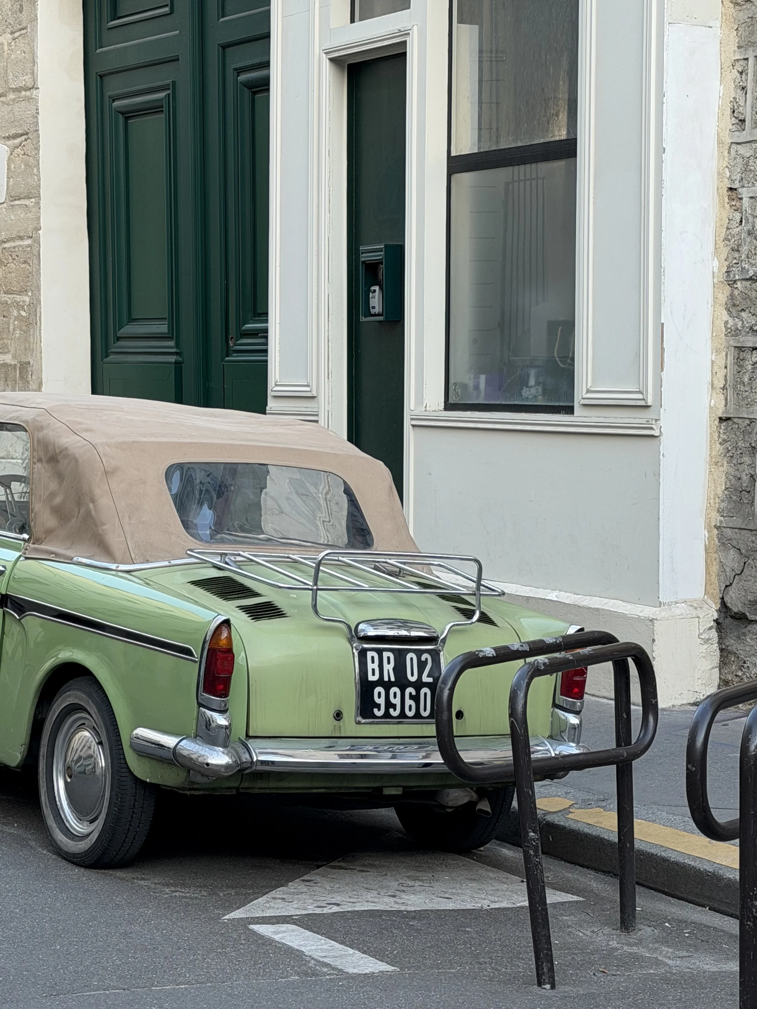 A vintage green convertible car parked on the street with a beige fabric roof, black and chrome bumper, and a black European license plate that reads BR 02 9960, next to a black metal bike rack in front of a building with a dark green door and a window.