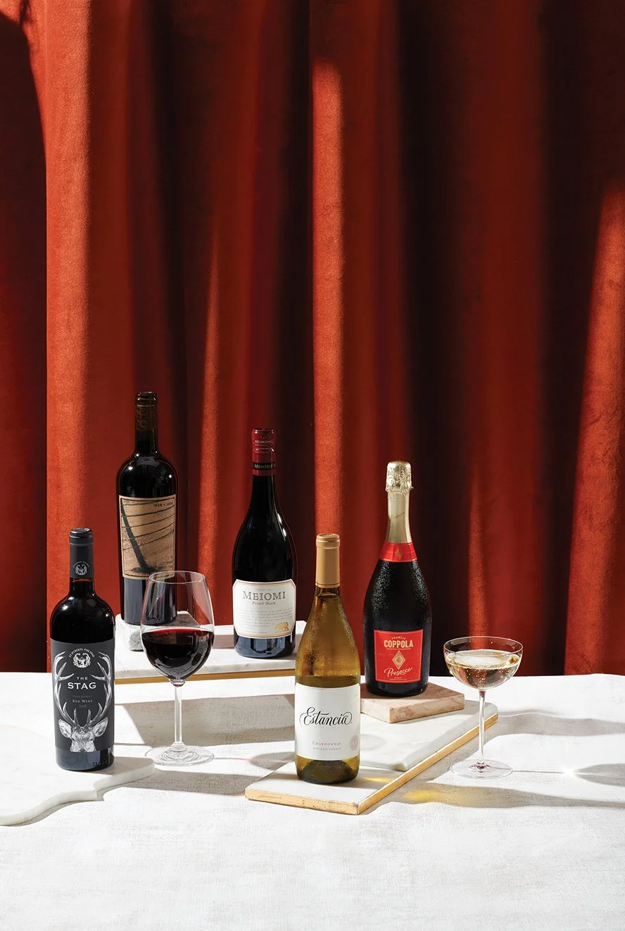 Selection of five bottles of wine and champagne with two glasses of red and white wine on a white table in front of a red curtain.