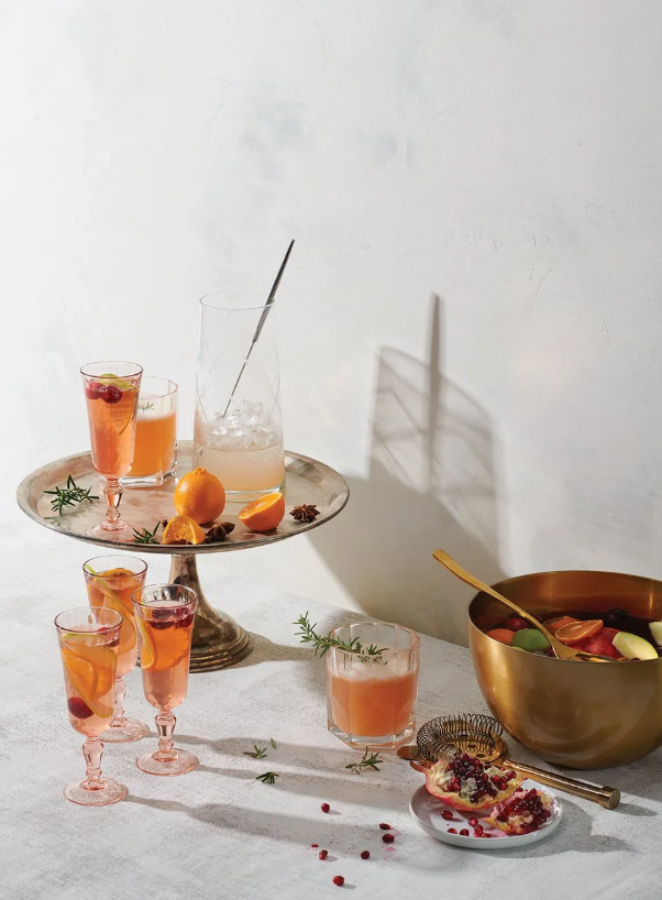 A table with a tiered tray holding pink and orange beverages, oranges with slices and star anise. A glass of pink drink, a bowl of fruit salad with apples, and a pomegranate with seeds and a gold spoon. The scene is lit with natural light casting shadows on the table and wall.