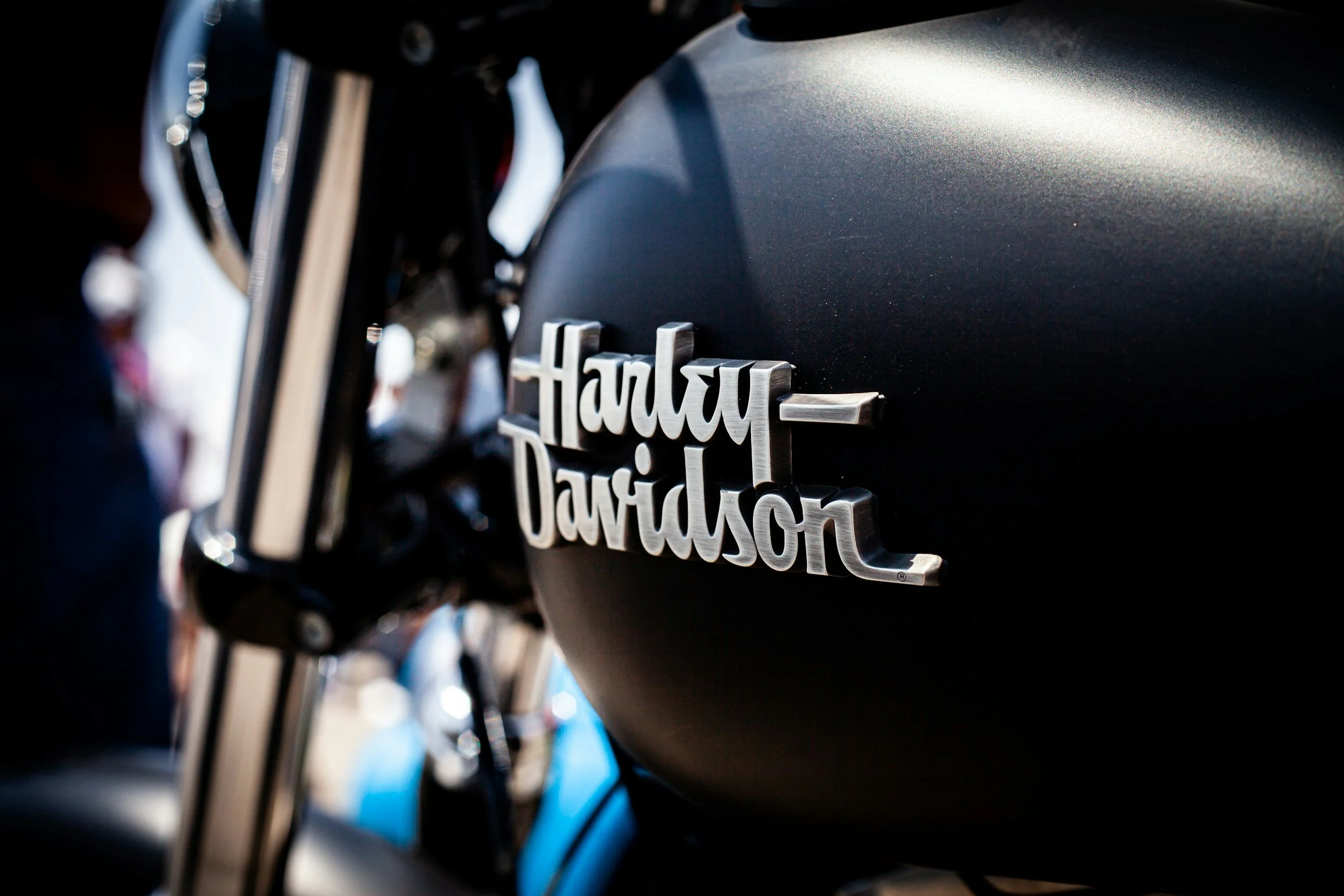 Close-up of a black Harley Davidson motorcycle with the Harley Davidson logo on the tank, with blurred people and bike parts in the background.