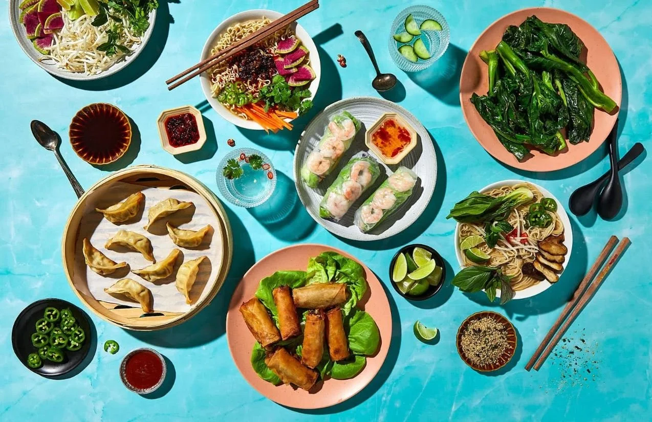 Variety of Asian dishes on a turquoise table, including spring rolls, dumplings, noodle salads, and fresh vegetables.