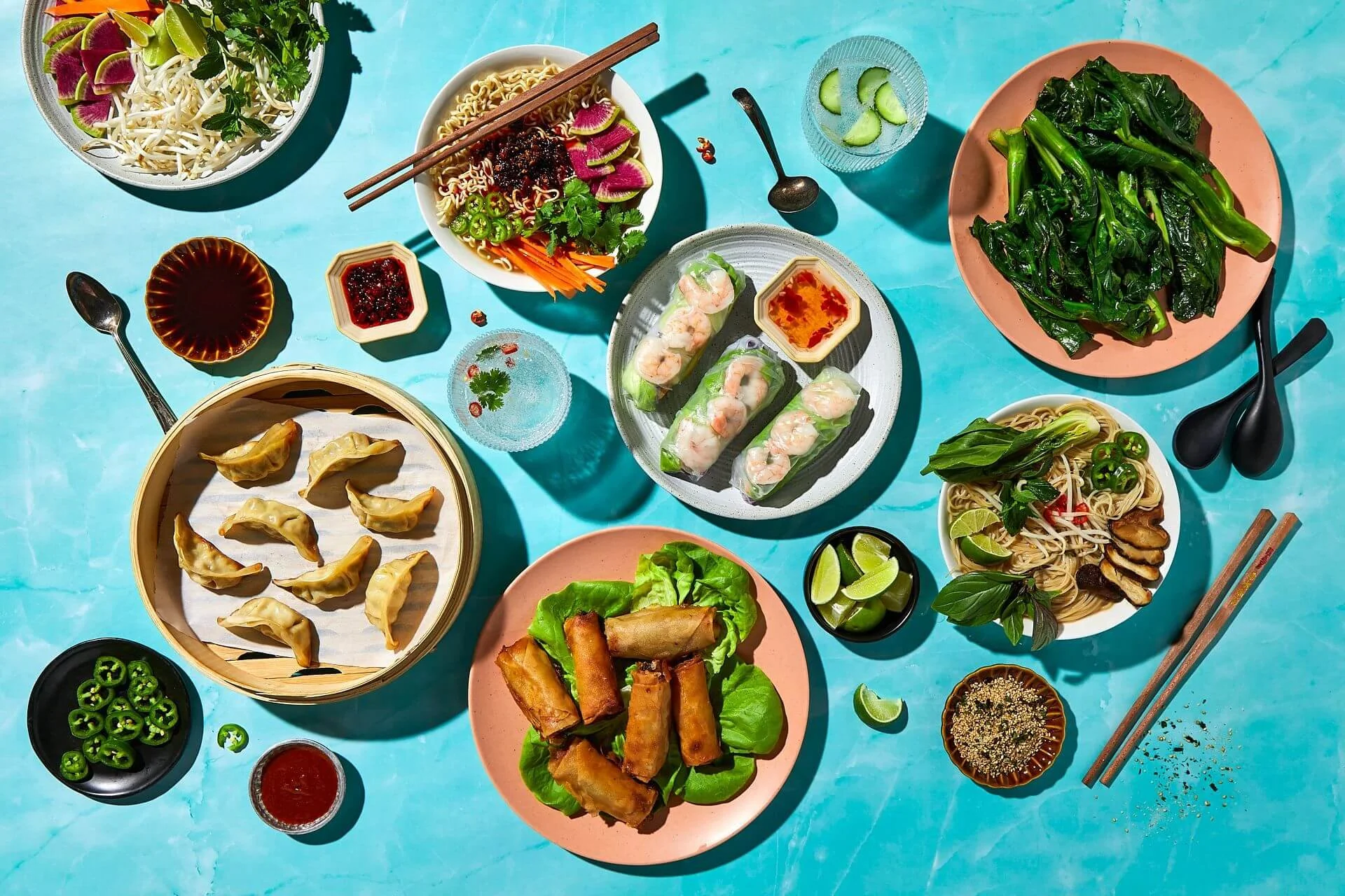 An overhead view of an Asian-style meal spread on a blue surface, featuring spring rolls, dumplings, noodles, greens, and various sauces and garnishes.
