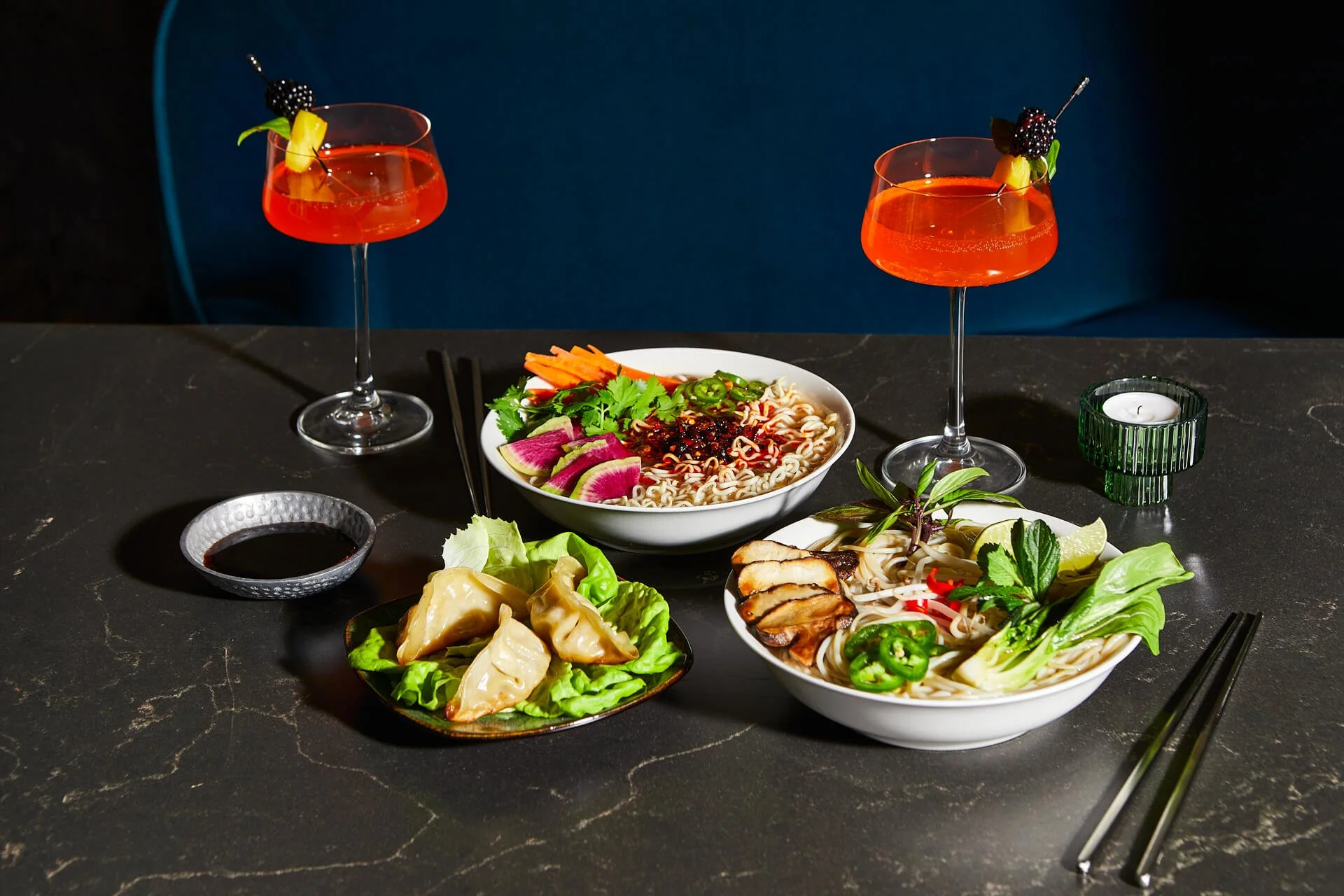 Two cocktails with garnishes, two bowls of ramen with vegetables and meat, a small plate of dumplings on lettuce, soy sauce, and chopsticks on a dark table