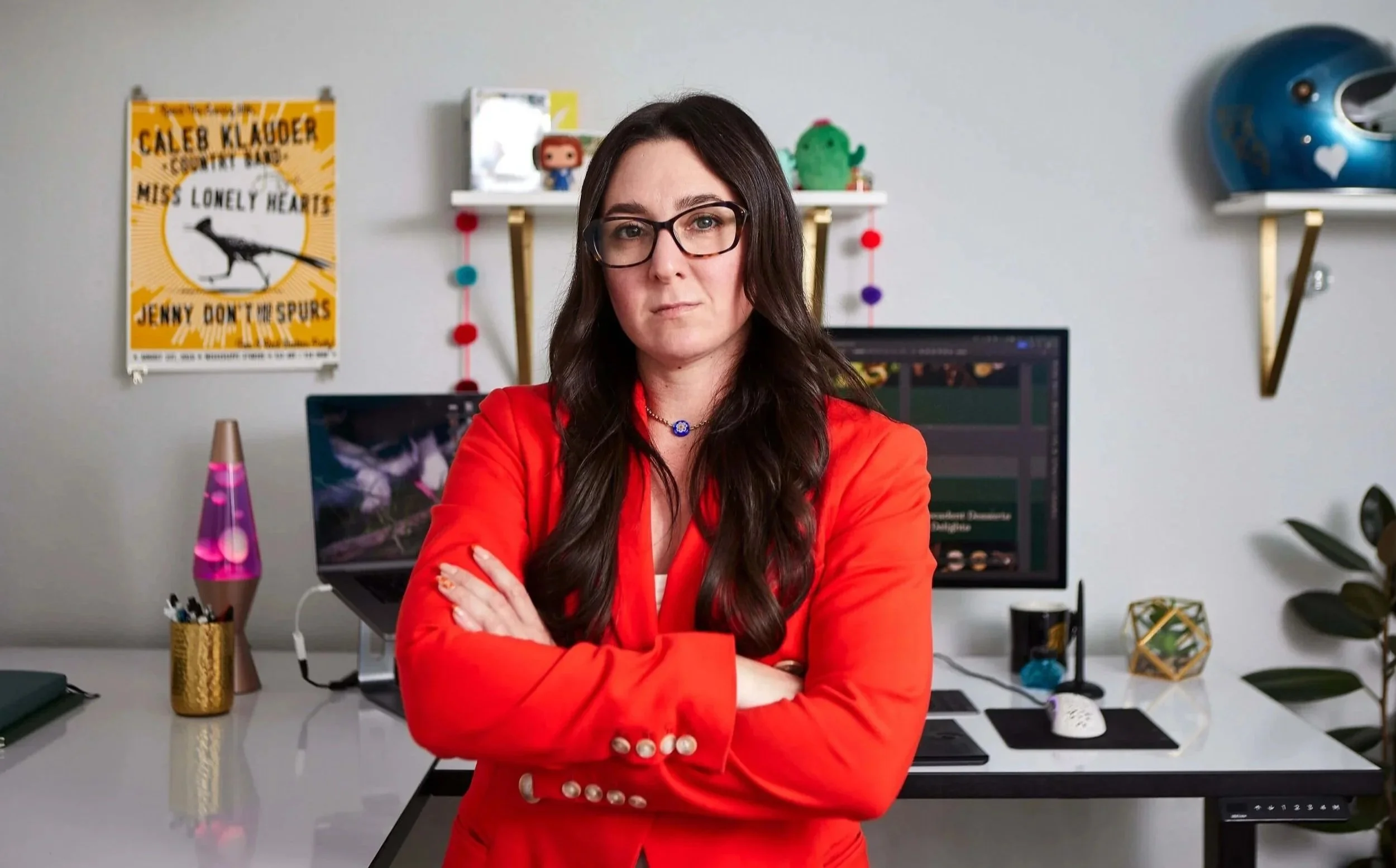 A woman with long dark hair and glasses wearing a red blazer crossing her arms in a modern office, with a computer, lava lamp, and shelves with decorative items and posters in the background.